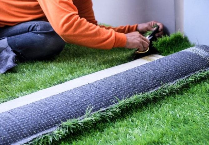 A person wearing an orange shirt trims the edges of artificial grass near a white wall with a pair of scissors.