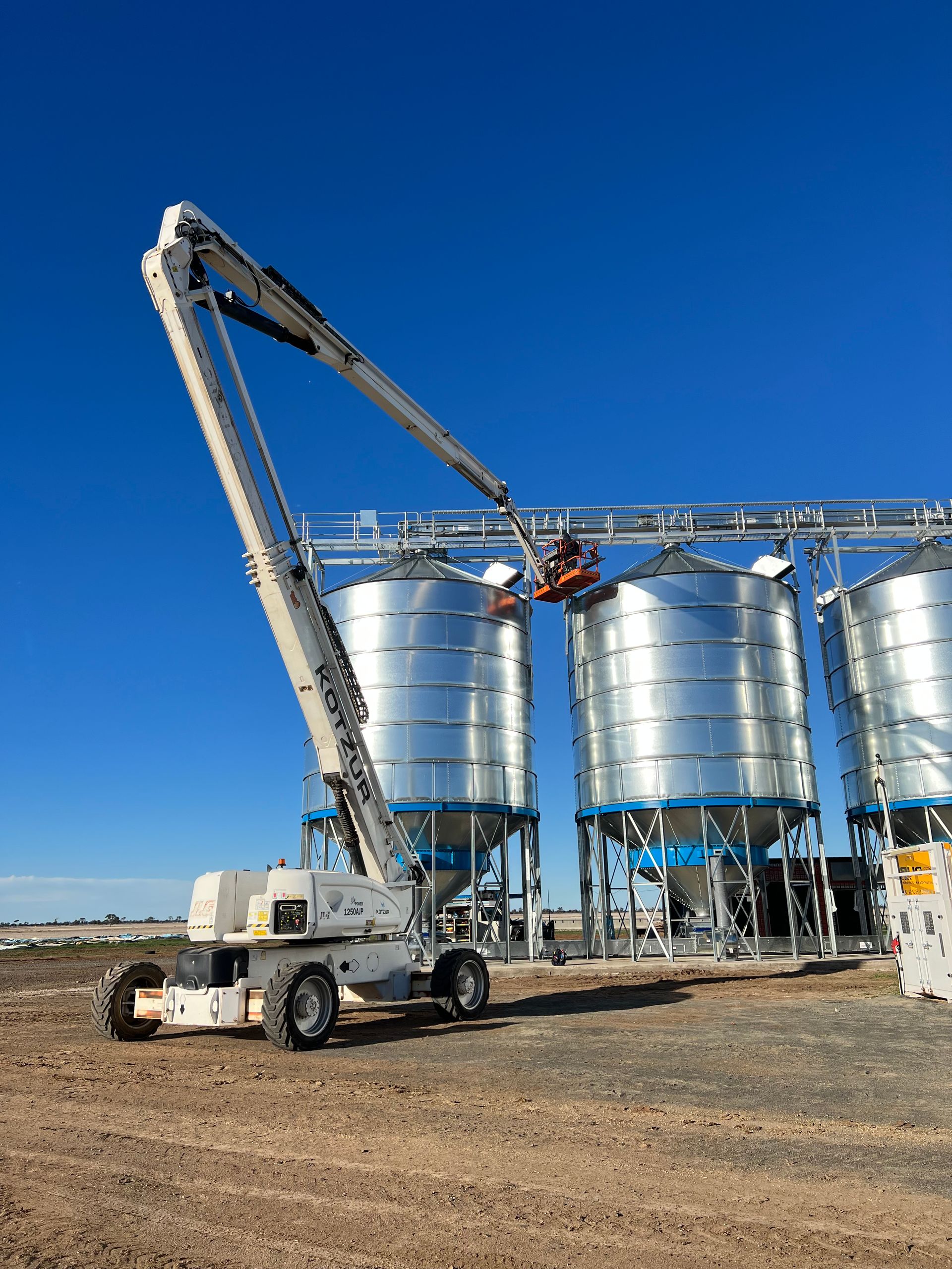 A Large White Material Handler Loads Grain Into Silver Silos Against a Bright Blue Sky — Rohr Electrical in Dubbo, NSW