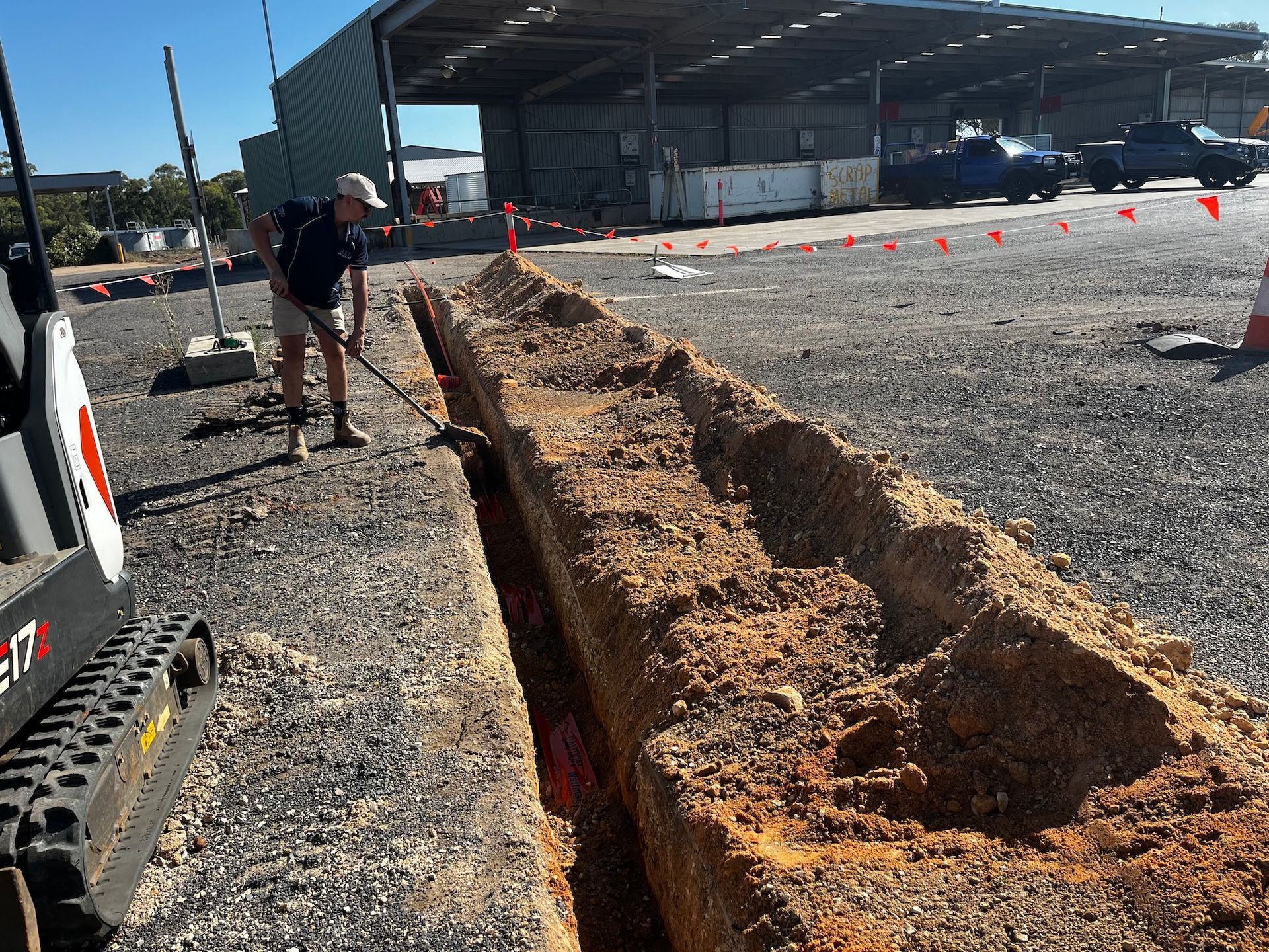 Man Shovels Soil From a Long Trench, With a Small Excavator Nearby — Rohr Electrical in Dubbo, NSW