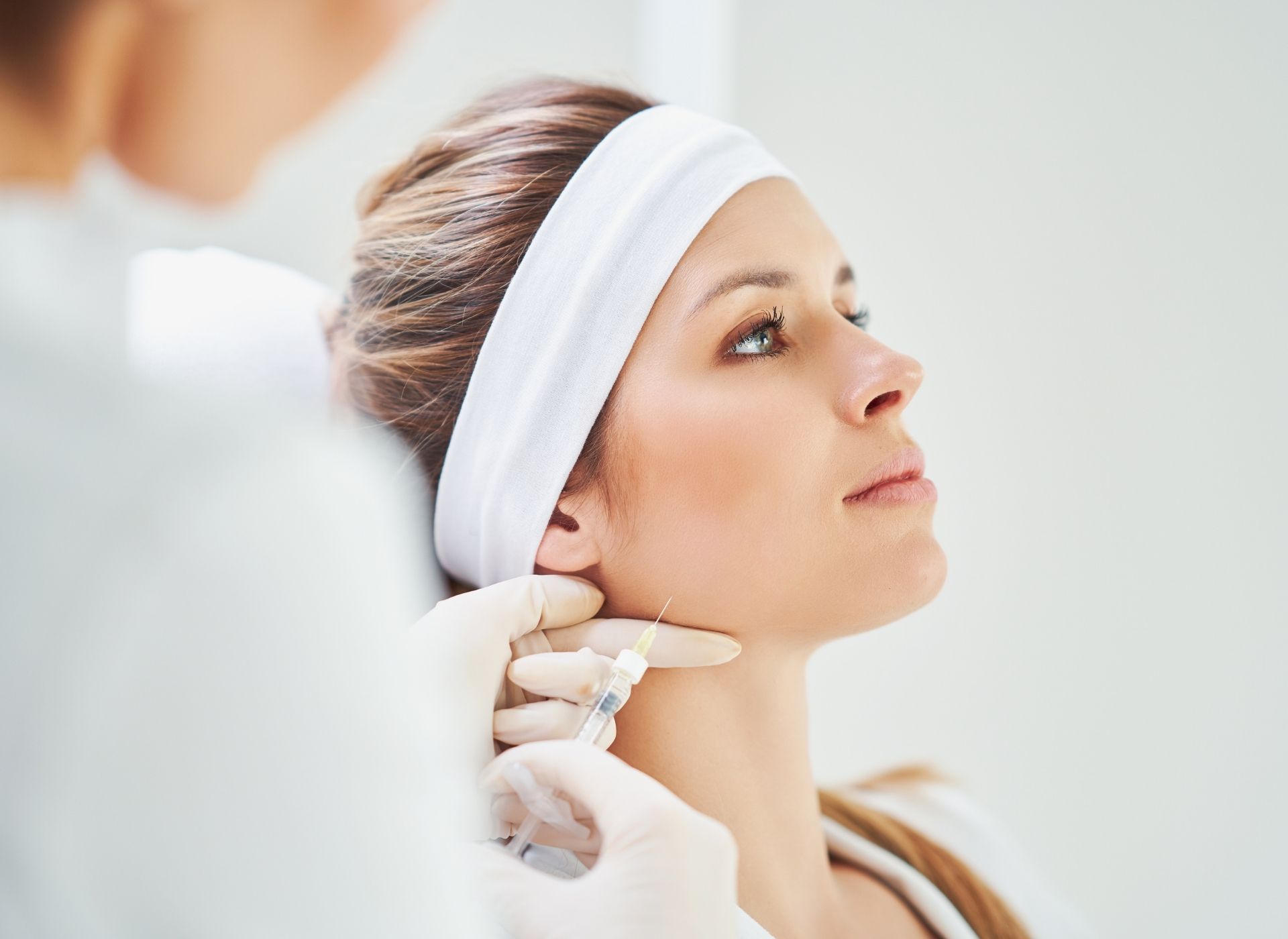 A person in a white headband receives a cosmetic injection in the neck from a medical professional wearing gloves.