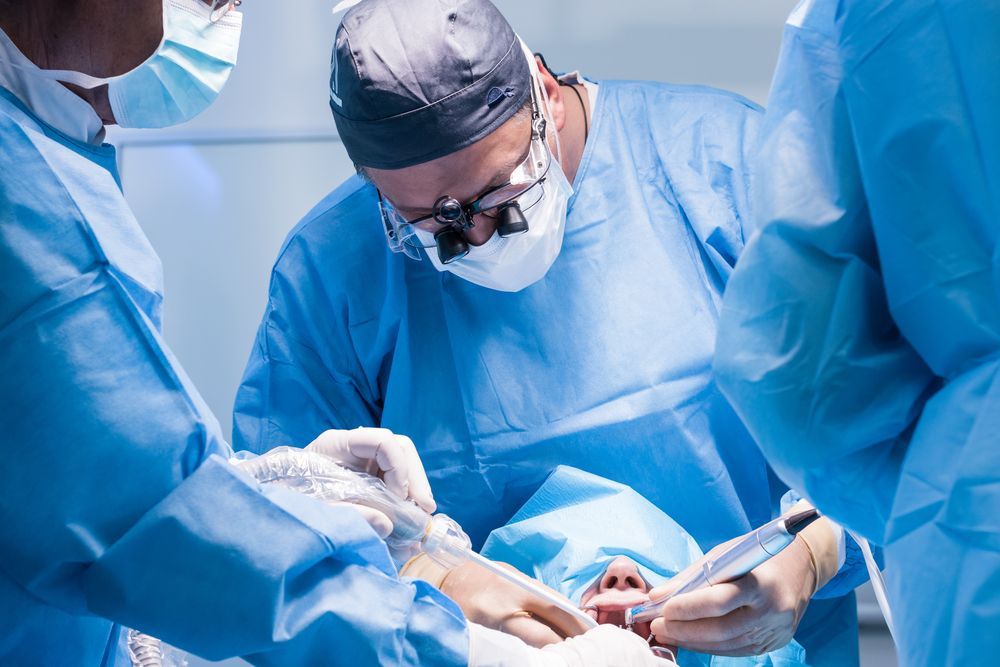 Surgeons in blue scrubs and surgical masks perform a dental procedure on a patient draped in a blue sterile cloth.
