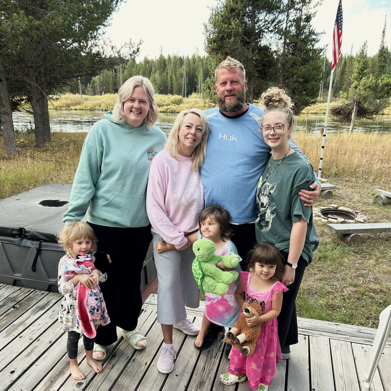 Family posing on wooden deck near water; adults and children smiling; American flag in background.