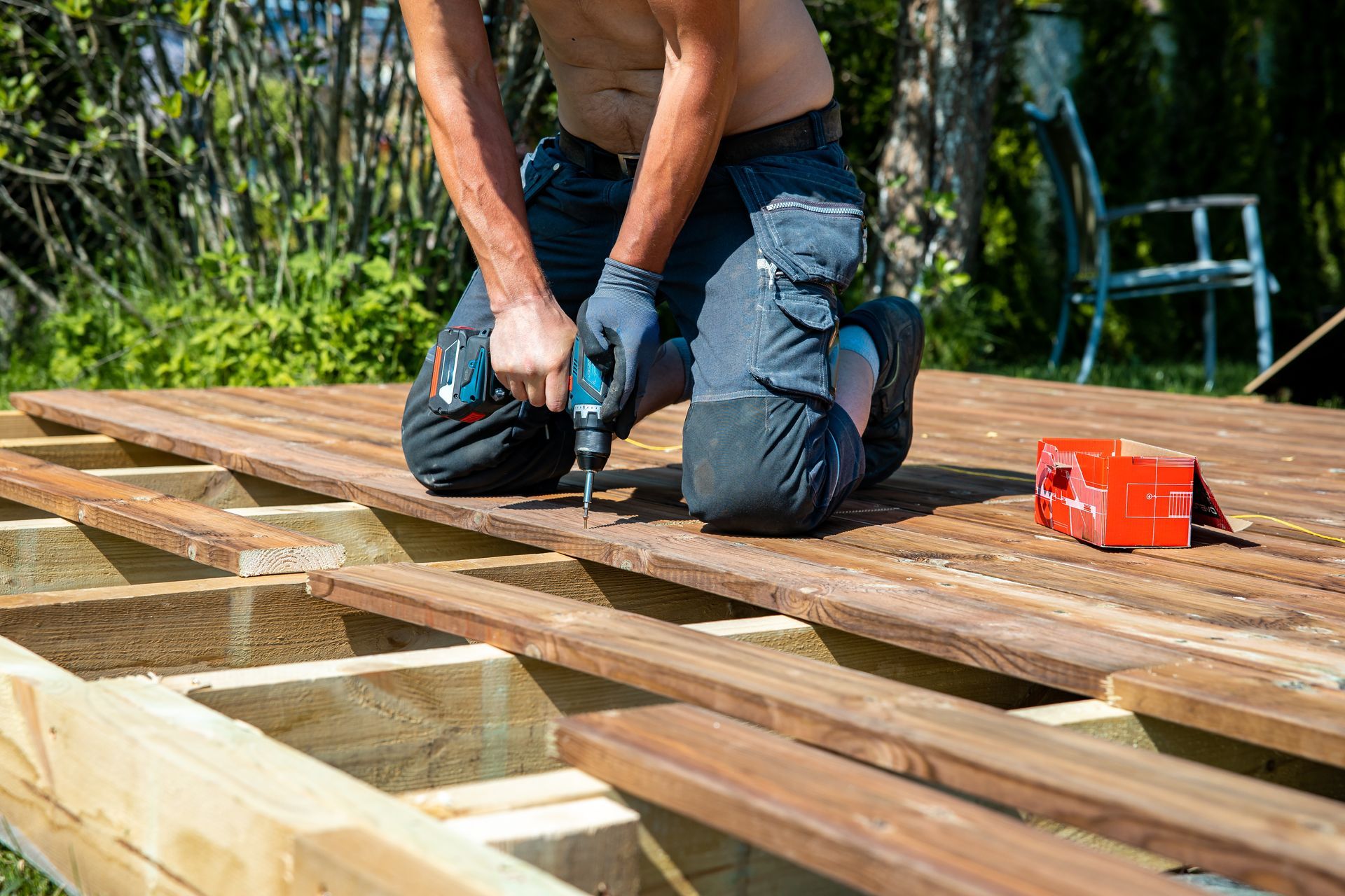 Worker kneeling on a wooden roof deck, using a nail gun beside a red toolbox outdoors