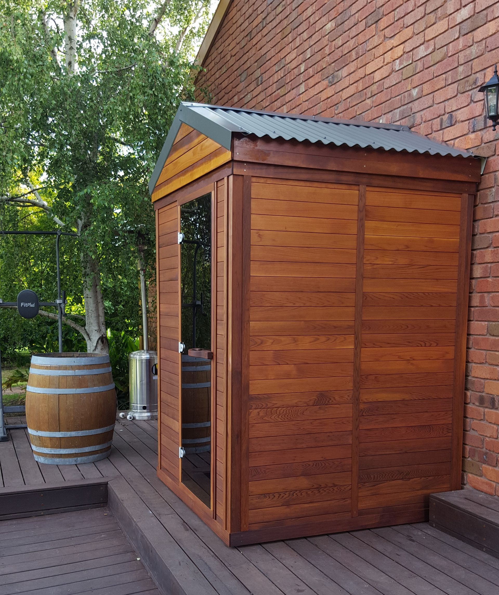 Wooden sauna shed with glass door beside a brick wall on a deck outdoors
