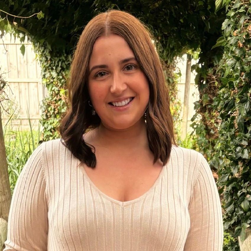 Smiling woman in a cream ribbed top standing outdoors among greenery and ivy.