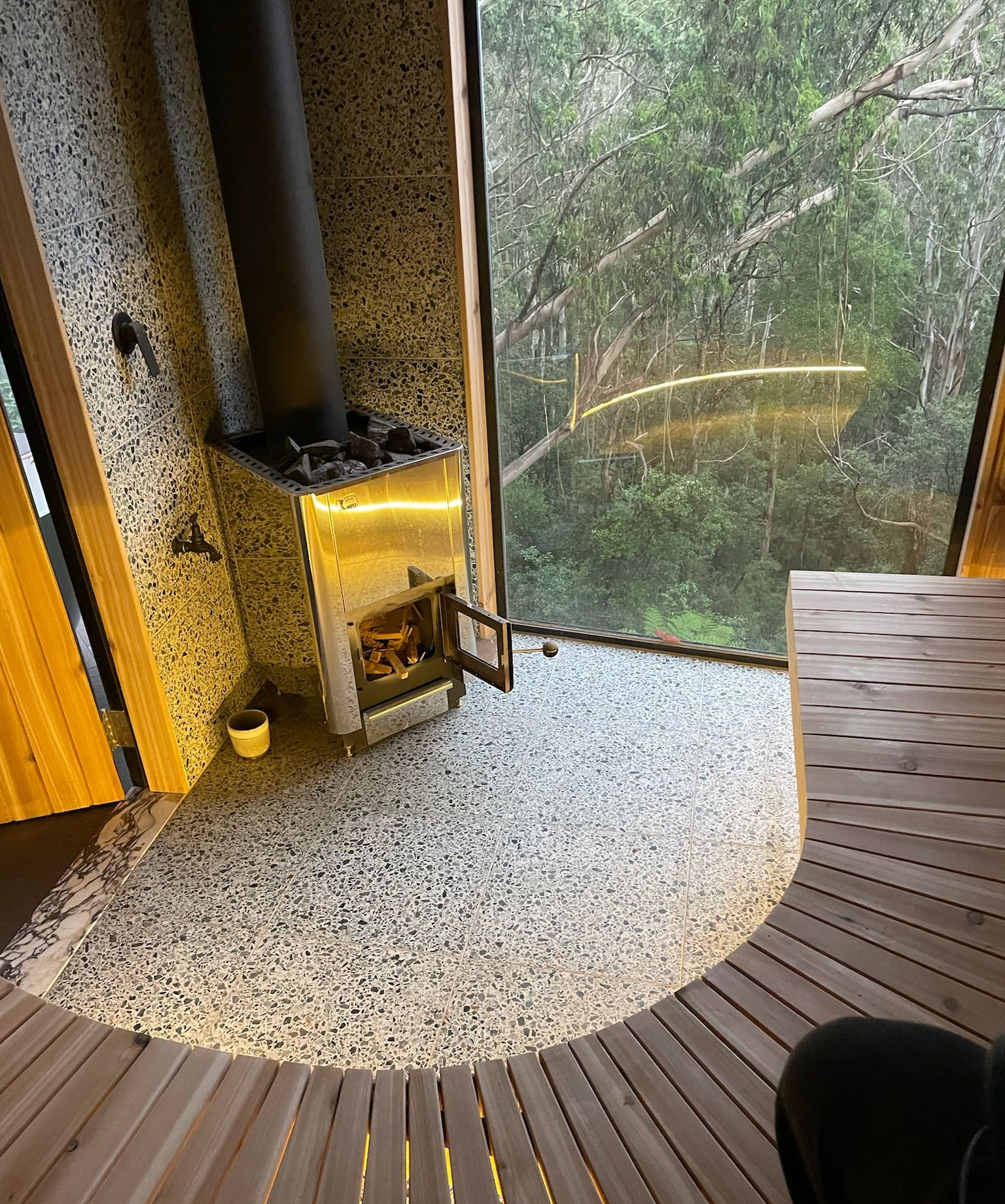 Curved indoor deck beside a small wood stove, looking out through floor-to-ceiling windows onto greenery
