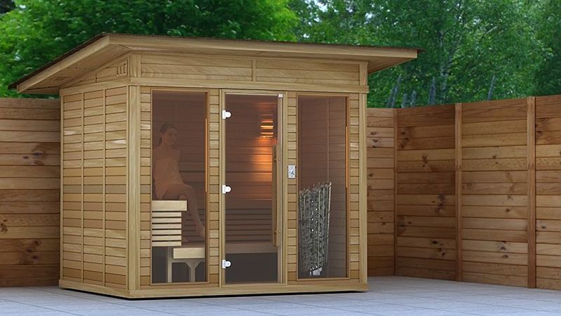 Small wooden sauna cabin with glass door and benches, set on a patio surrounded by a fence and trees.