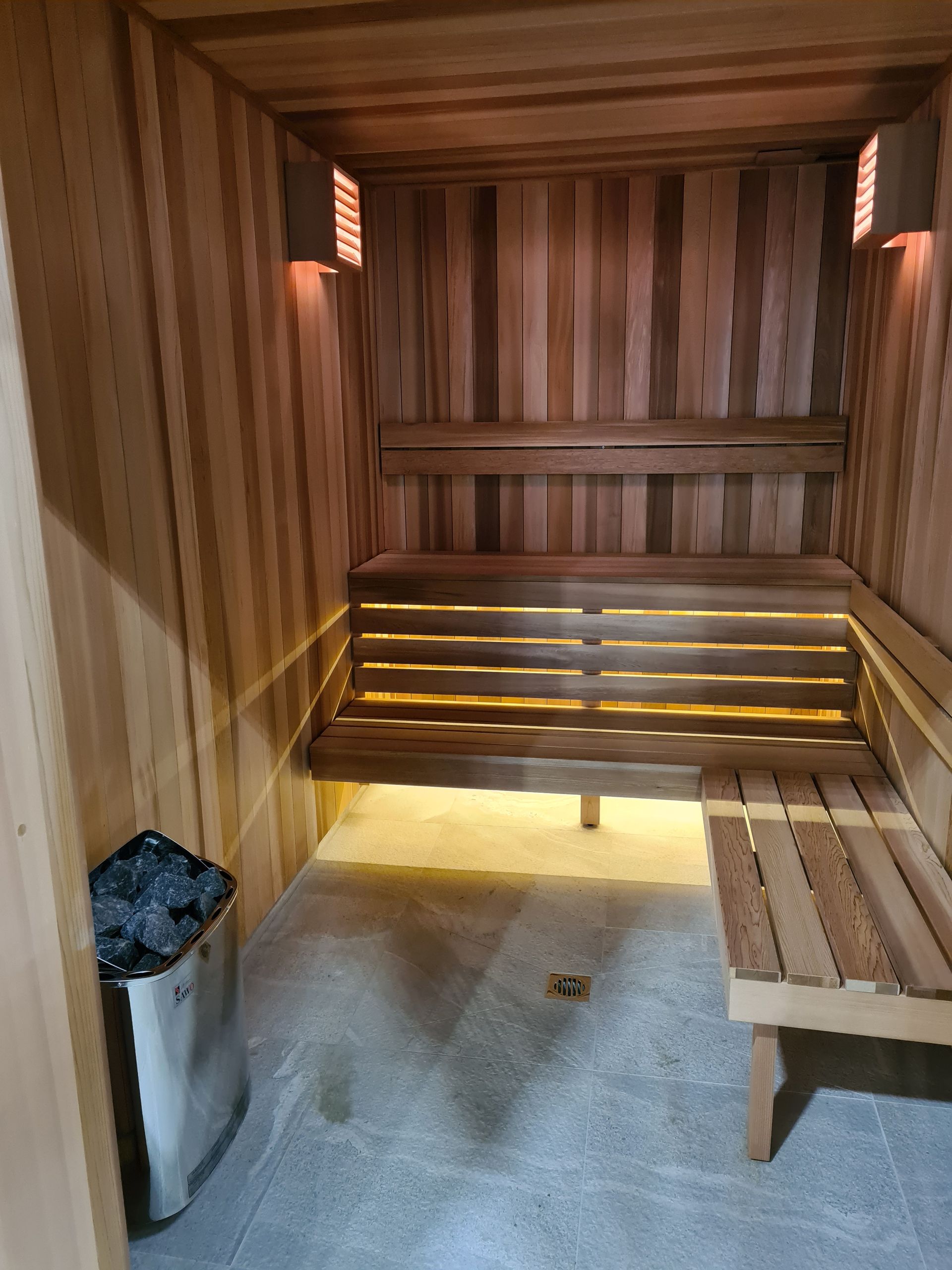 Wood-paneled sauna with benches, warm lighting, and a small trash bin by the door.