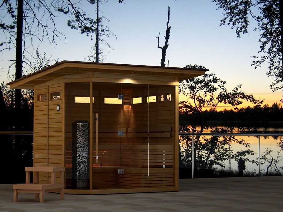 Small wooden lakeside sauna at sunset with warm lights and a bench on the deck
