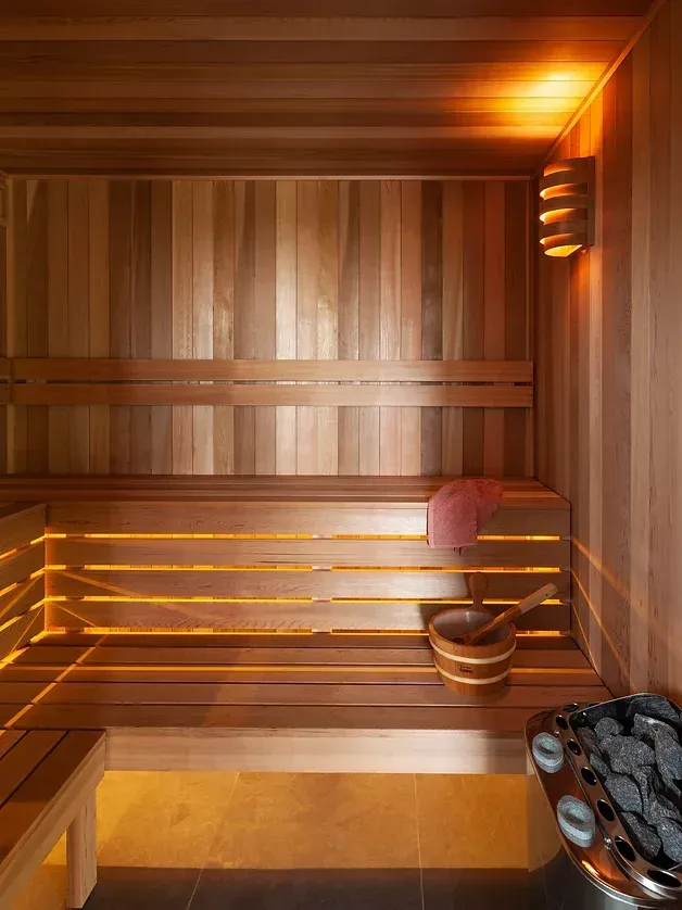 Wood-paneled sauna with benches, warm orange lighting, and a bucket and ladle on the floor