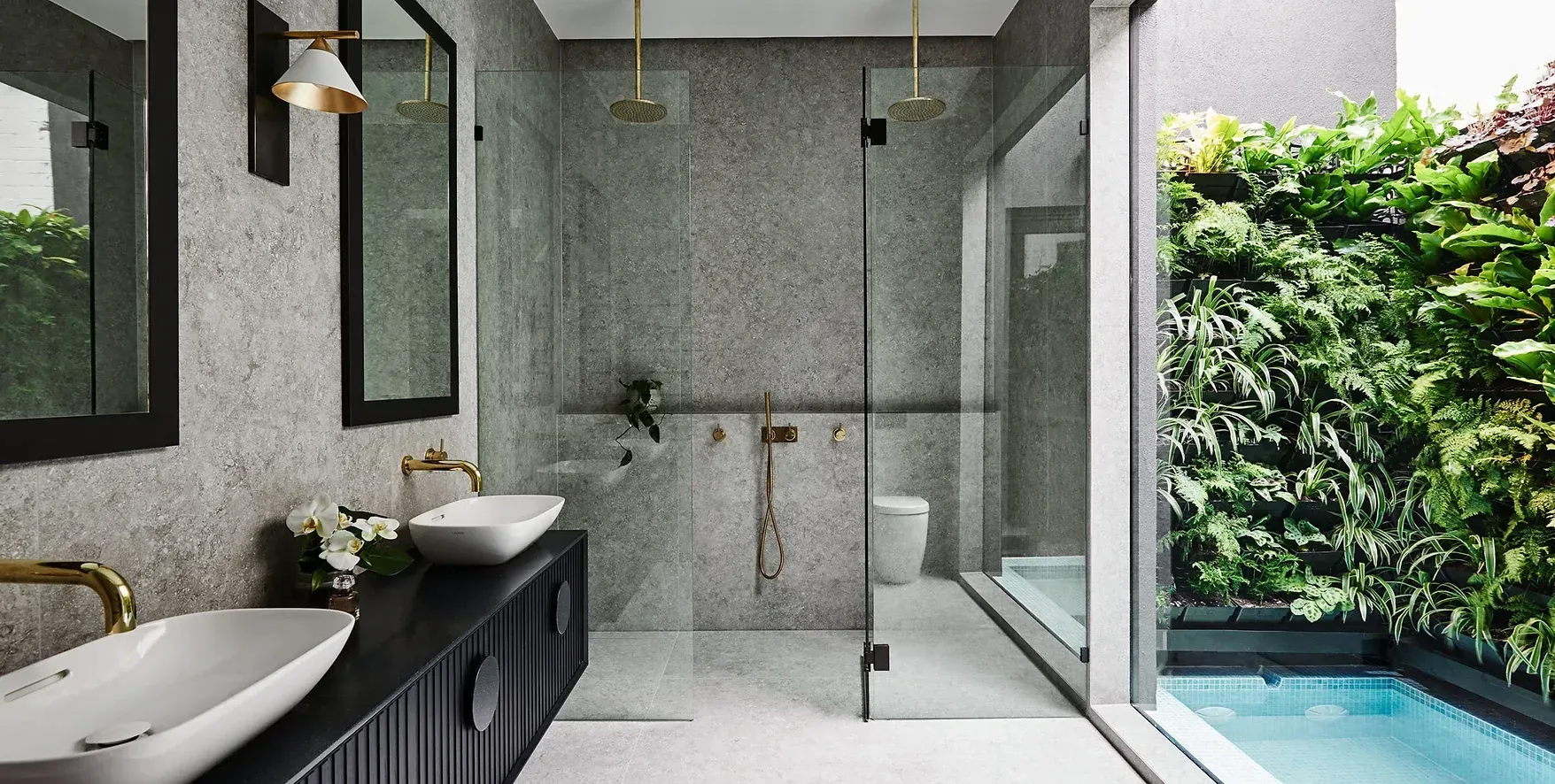 Modern bathroom with double sinks, glass shower, and a view of lush greenery and a pool outside