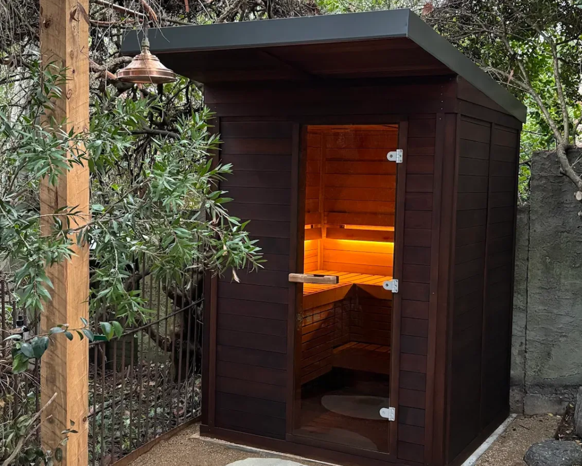 Small dark wooden outdoor sauna with an open door and warm orange light inside, beside greenery.