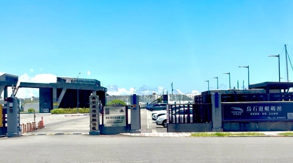 陽光明媚的日子，來到一個設施入口，那裡有金屬大門、停放的汽車和藍色牆上的標誌。