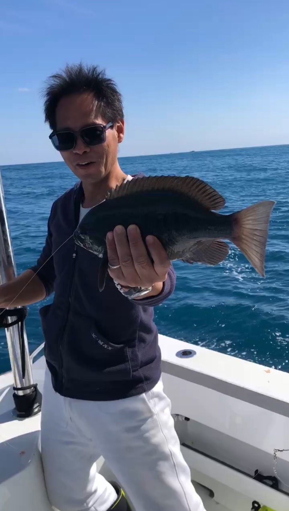 一名戴著太陽眼鏡的人站在船甲板上,拿著一條深色的魚對著鏡頭,背景是大海。