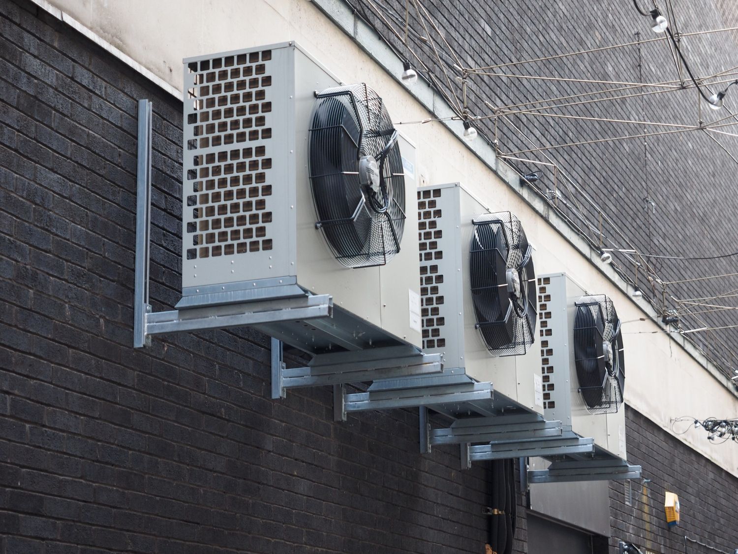 Three wall-mounted outdoor air conditioner units on a brick building under an overhang.