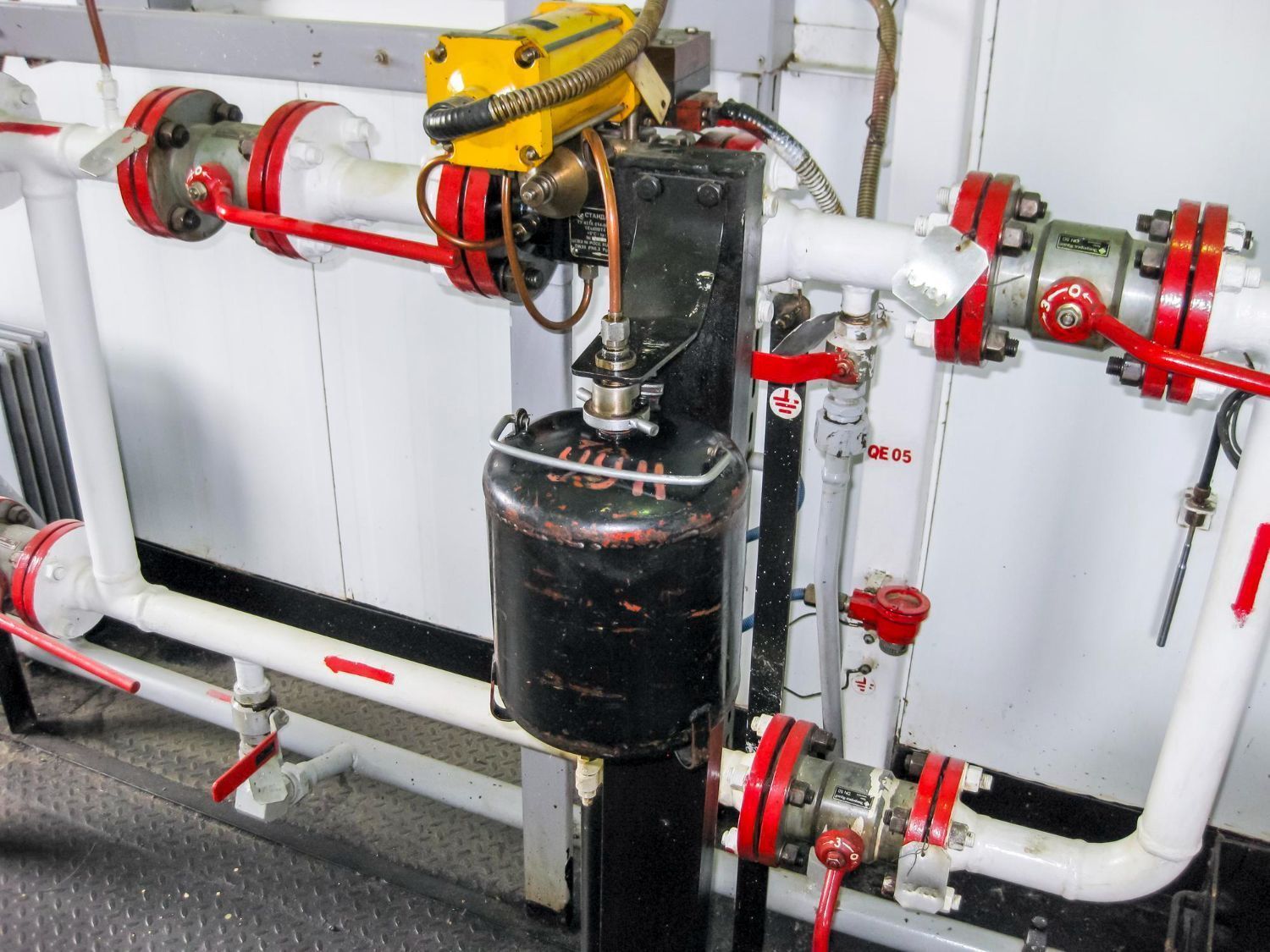 Industrial piping and valves with a black tank and yellow actuator in a mechanical room