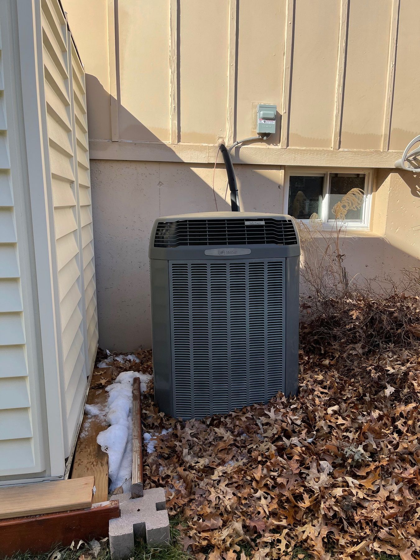 Outdoor HVAC unit beside a beige house, with patches of snow and brown shrubs nearby