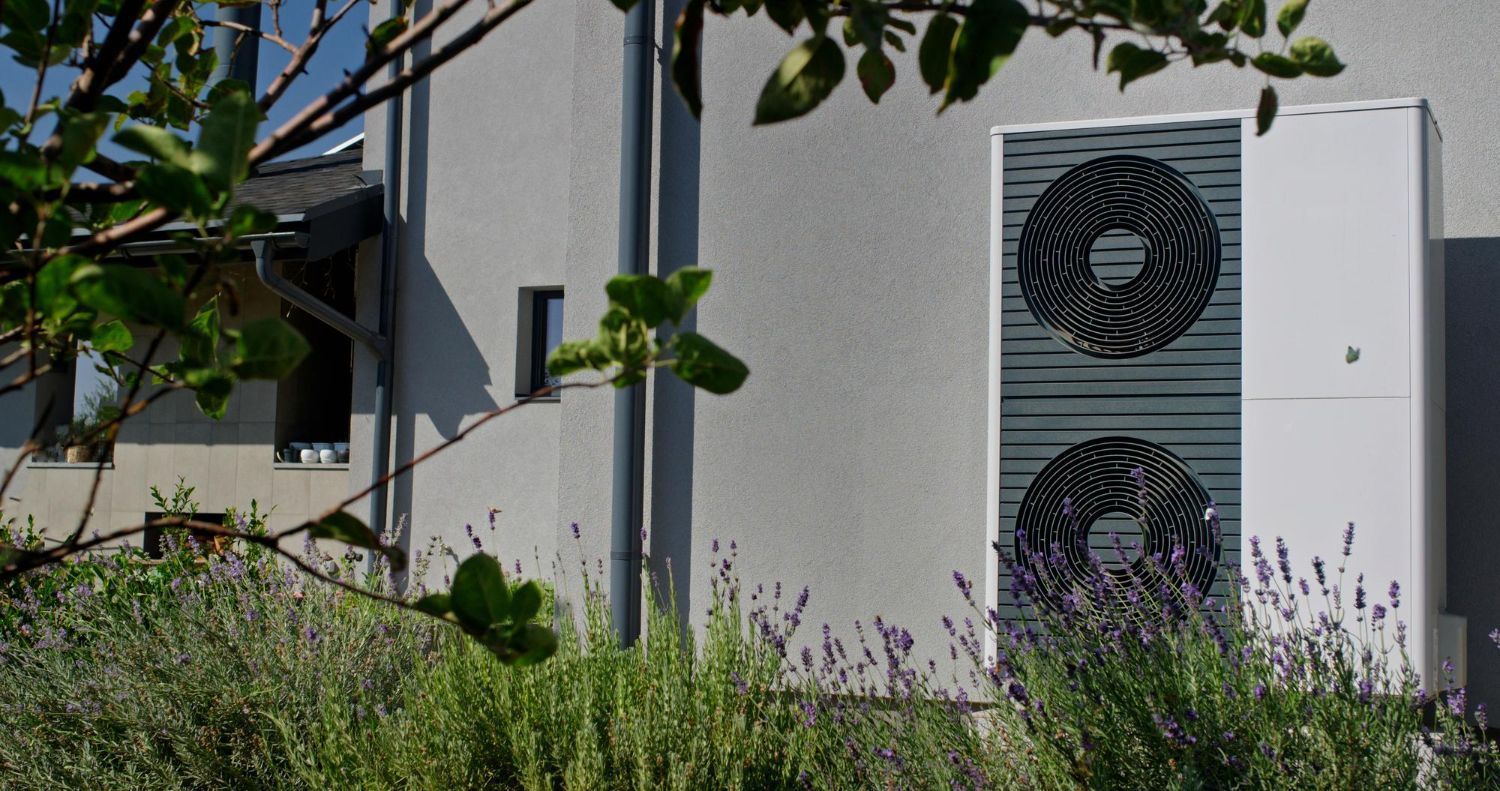 Outdoor air conditioner unit beside a house wall, partly obscured by green plants.