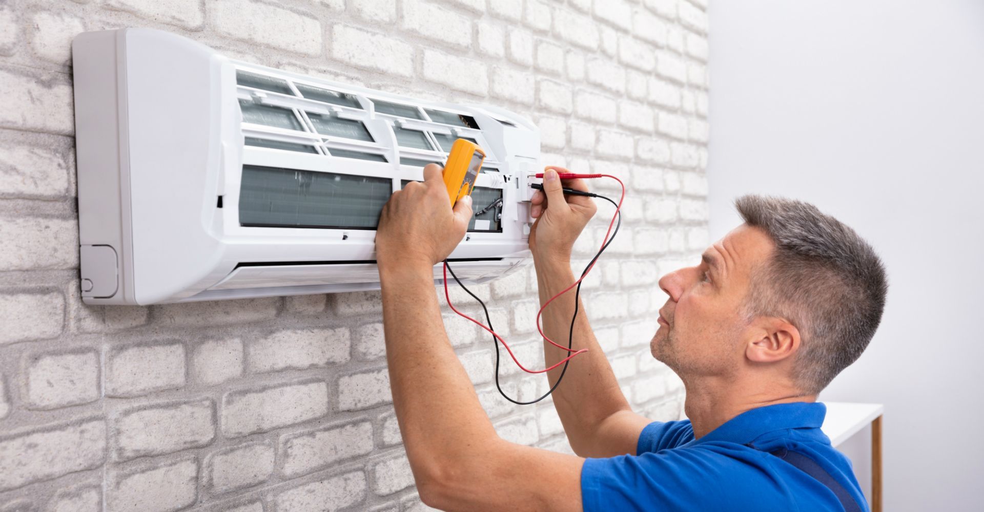 Technician testing an air conditioner on a brick wall with a screwdriver and multimeter
