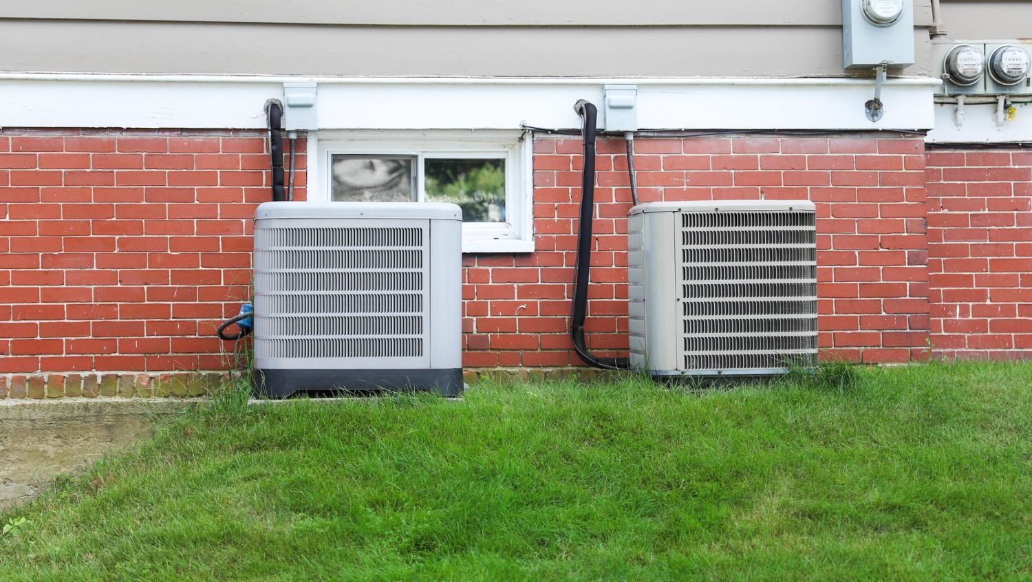 Two large gray HVAC units beside a brick building with a small window and grassy lawn.