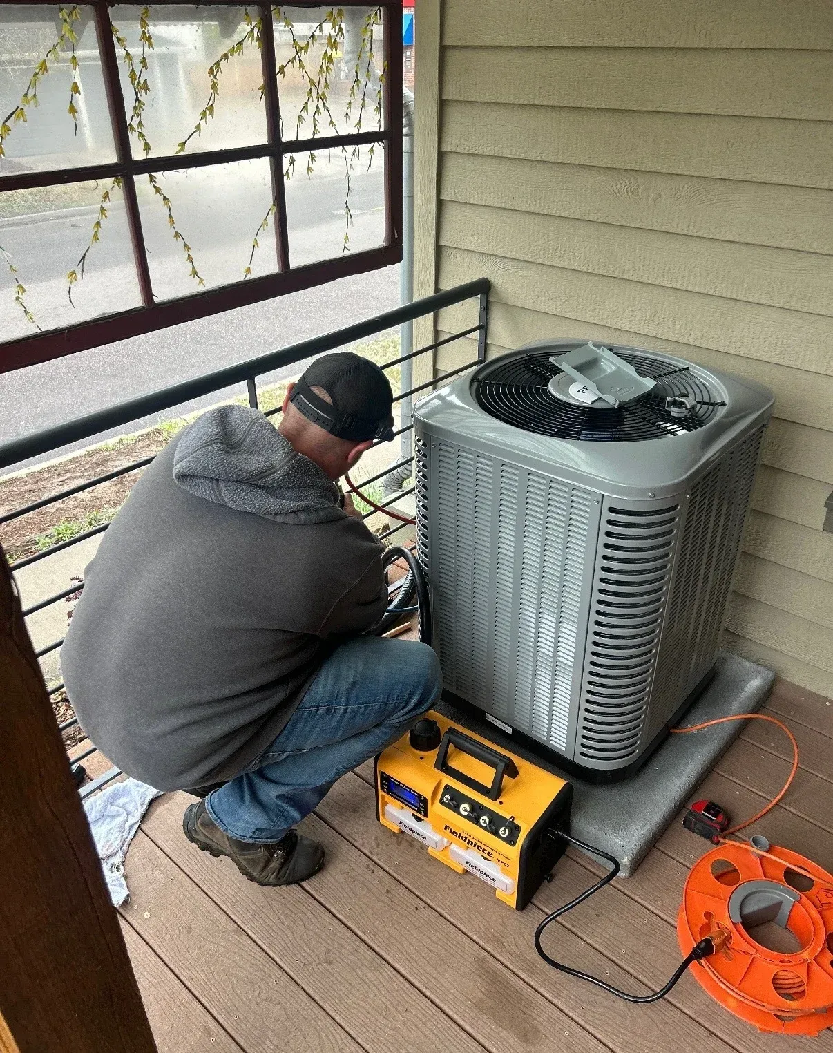 Technician kneels beside an outdoor HVAC unit, using tools on a covered porch.