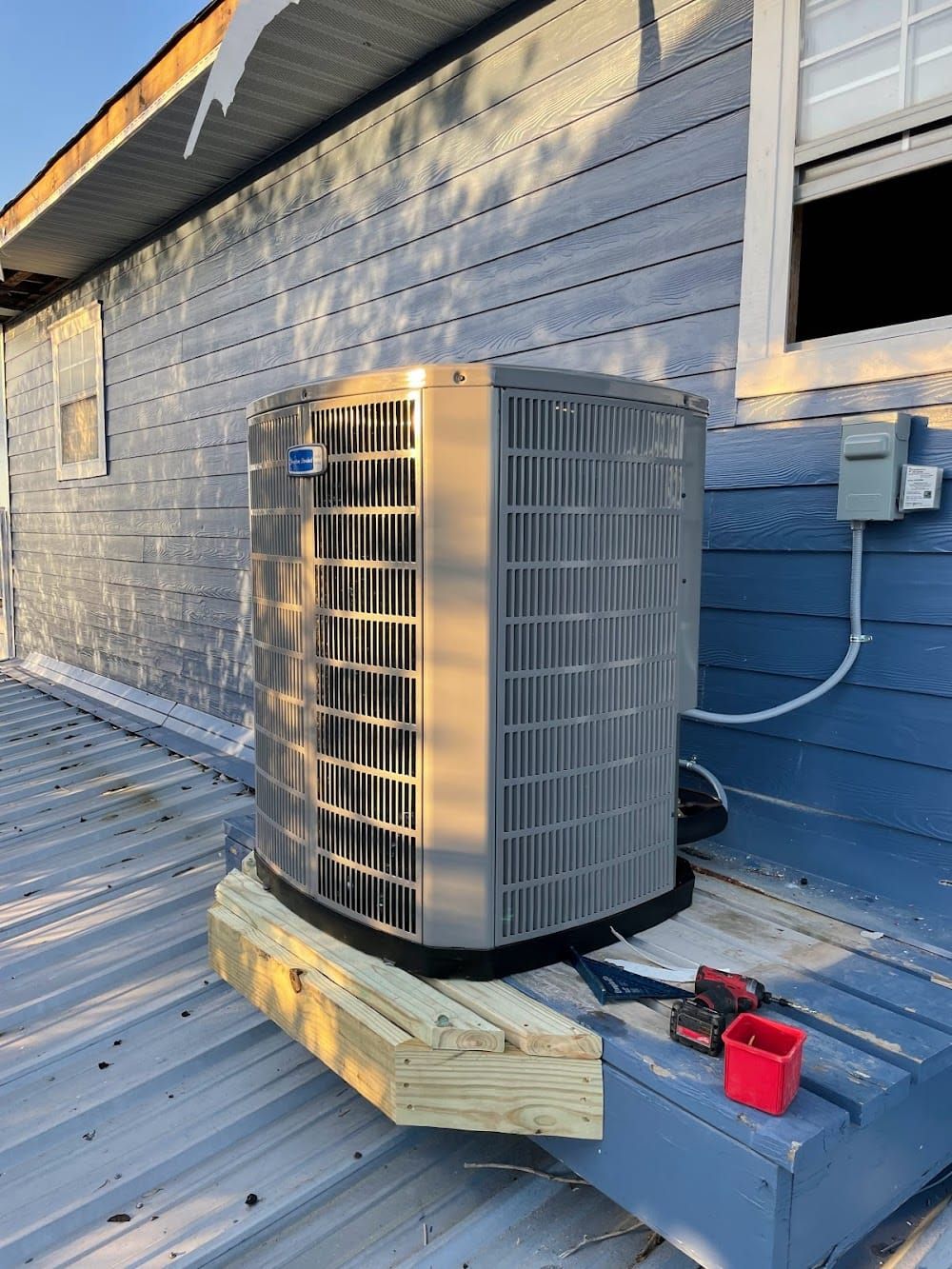 Outdoor HVAC condenser unit on a roof beside a blue wall and an open window, with a red tool nearby