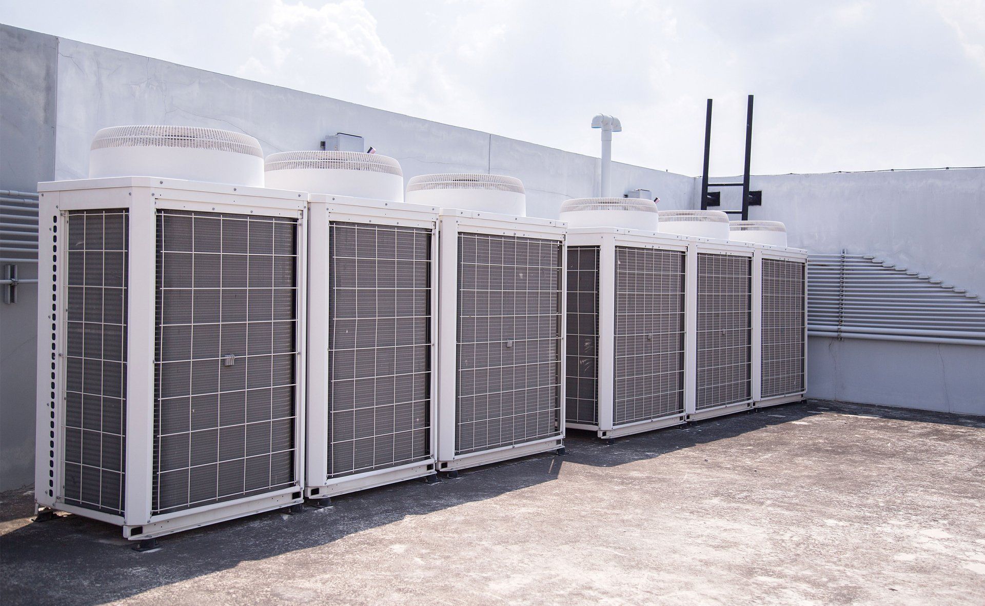 Row of outdoor HVAC units with metal grilles on a rooftop, beside a white wall