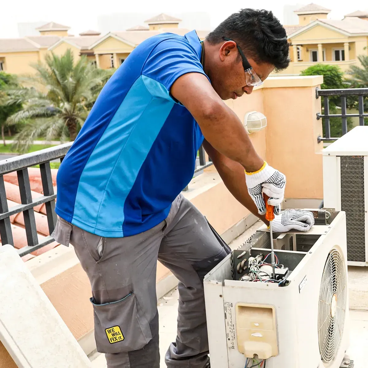 Technician in blue shirt repairing an outdoor air conditioning unit on a balcony