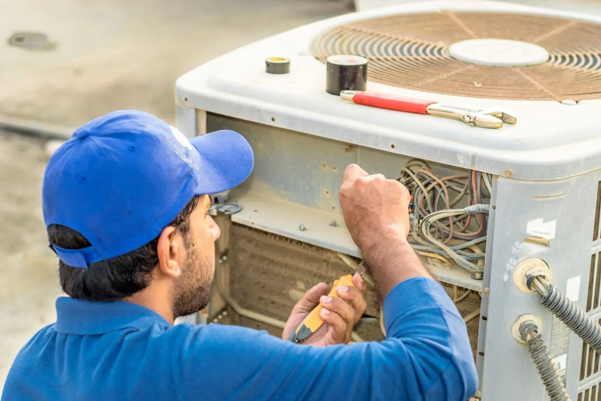 Technician in blue cap repairs outdoor air conditioner unit with exposed wiring