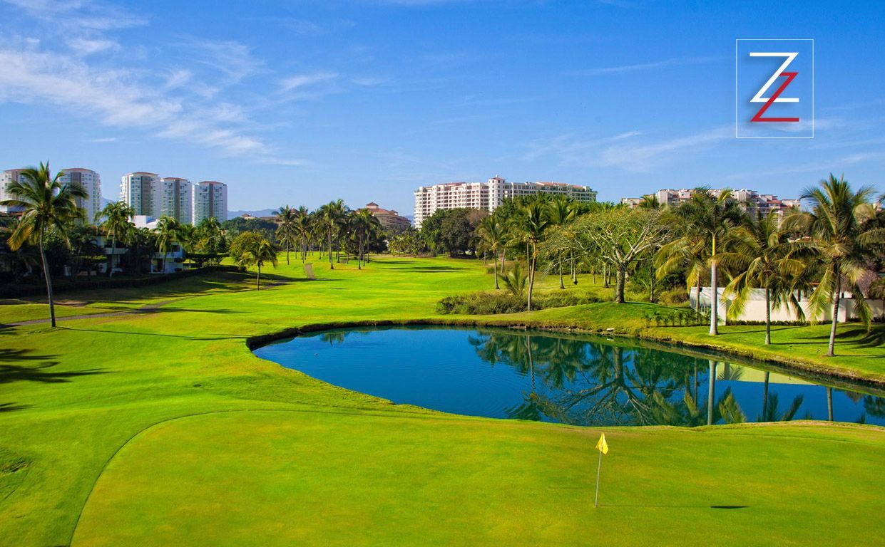 A golf course green with a small pond in the foreground and apartment buildings in the background under a blue sky.