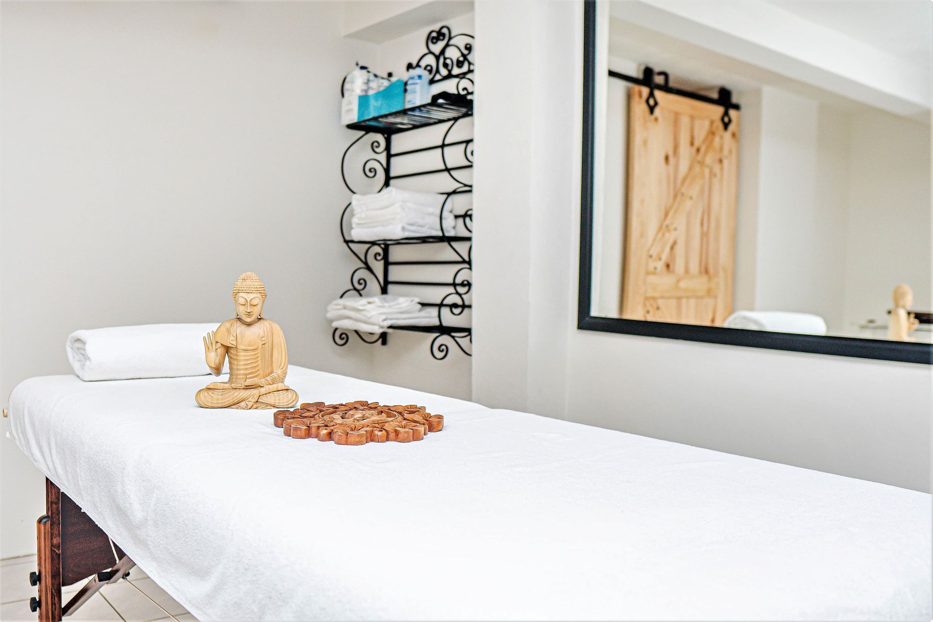 A massage table with a buddha statue and decorative stones, set in a clean, minimalist room with a towel rack and mirror.