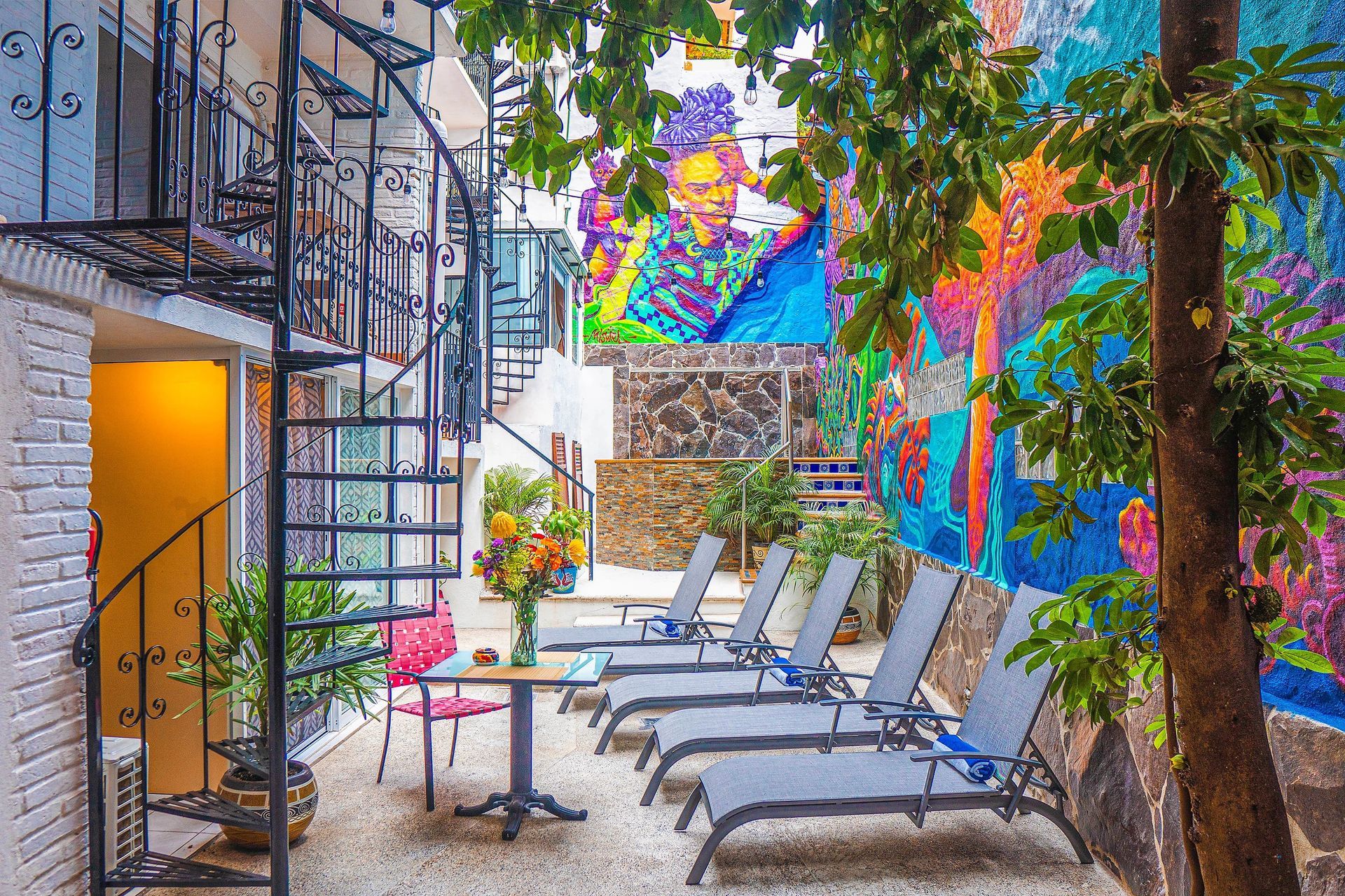 Courtyard with lounge chairs and colorful mural, spiral staircase, and potted plants.