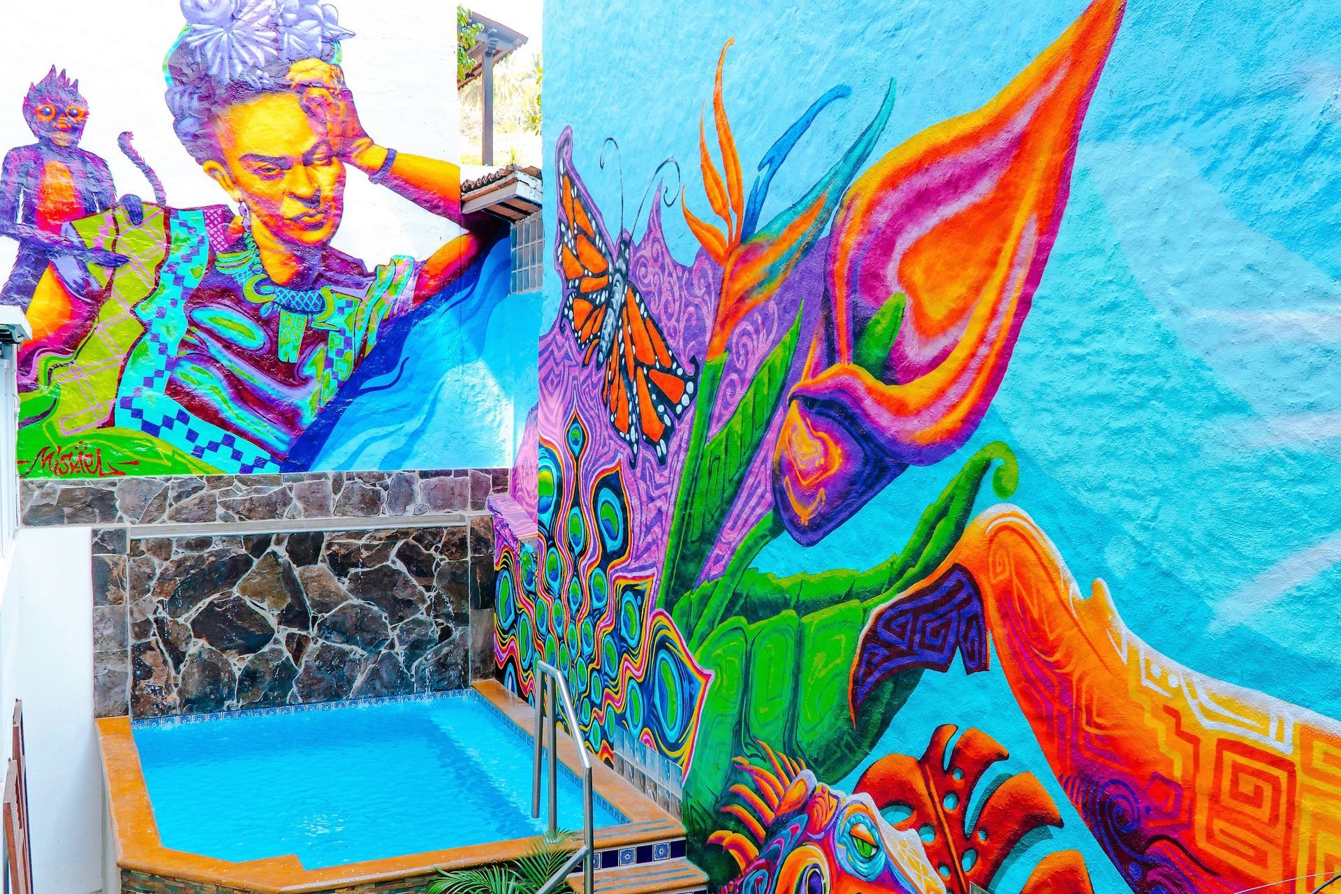 Colorful mural with Frida Kahlo portrait and abstract floral art near a small pool.