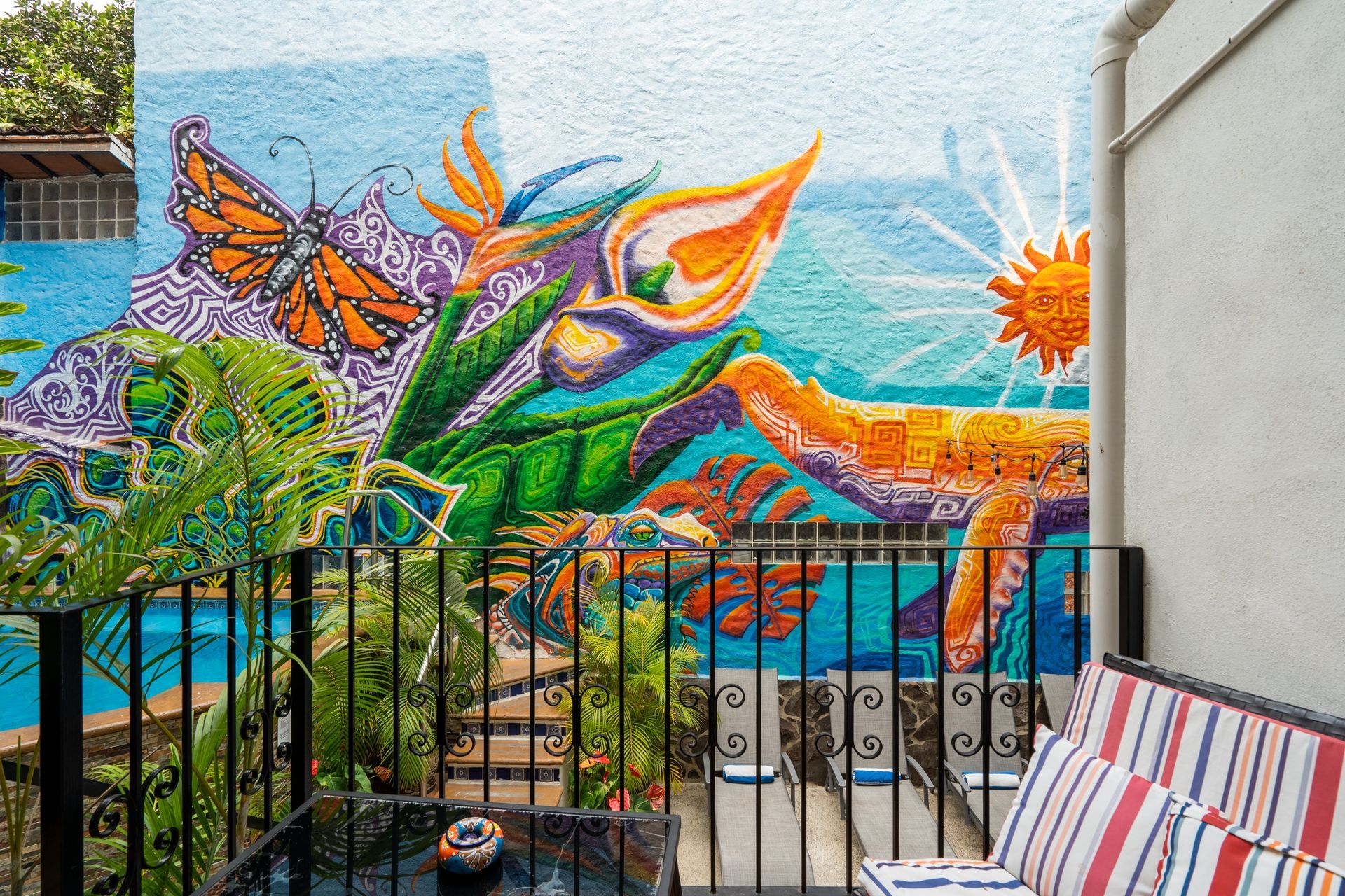 Mural of a monarch butterfly, calla lilies, and sun on a blue wall; view from a balcony with seating and a black table.