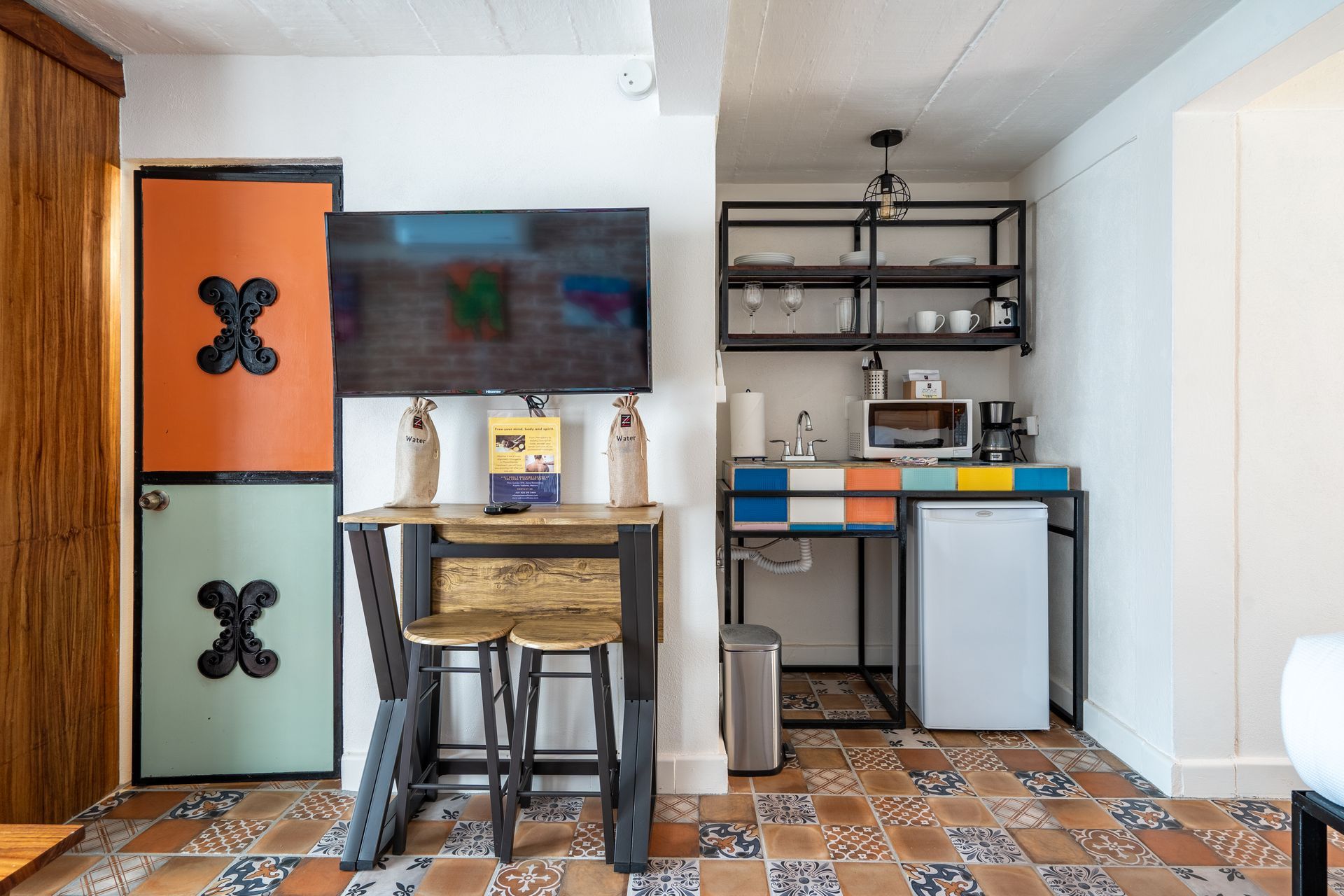 Small kitchen area with mini-fridge, shelves, microwave, sink, and table with stools in a room with patterned tiles.