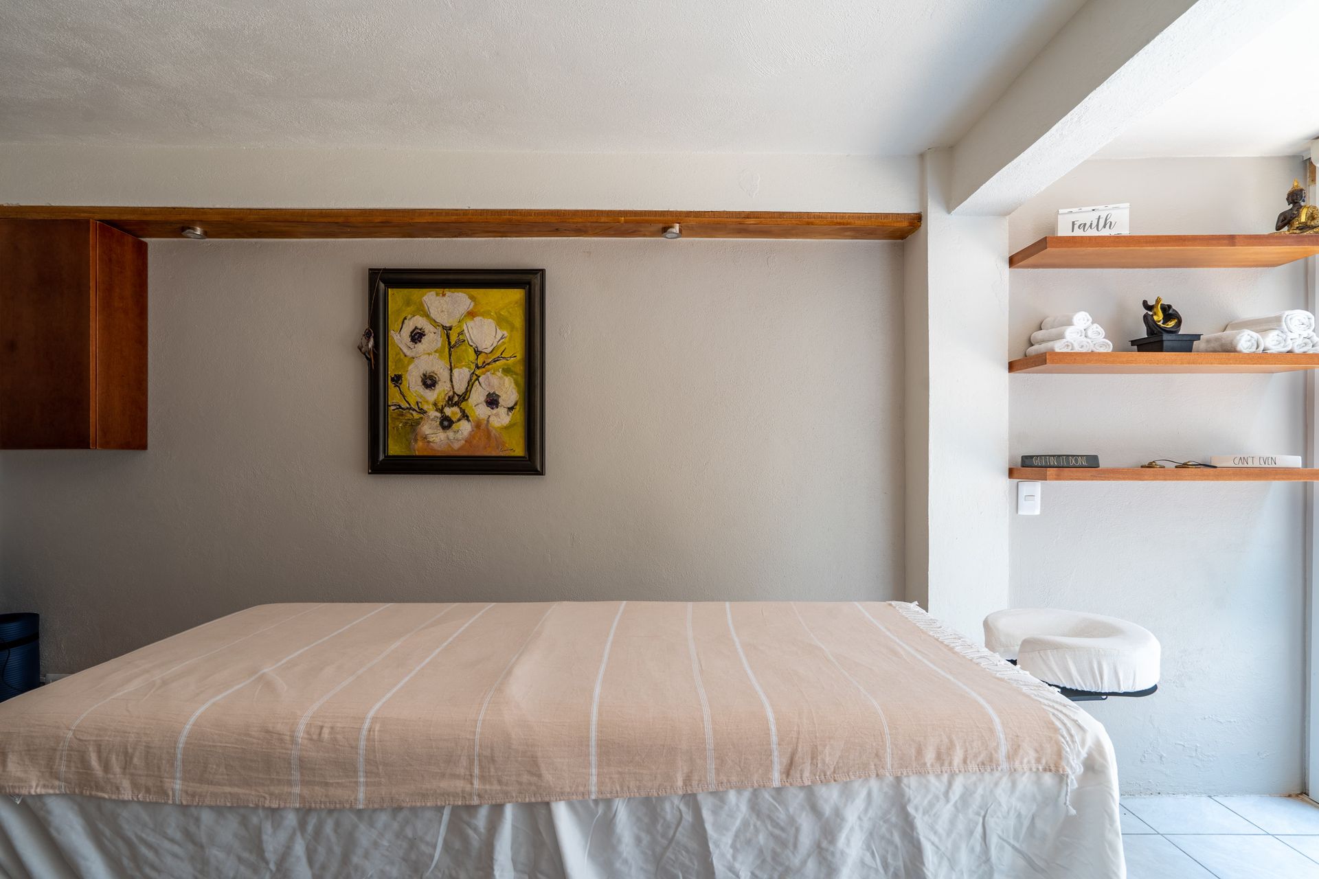 A massage therapy room with a covered massage table, wall art, and open wooden shelving holding supplies.