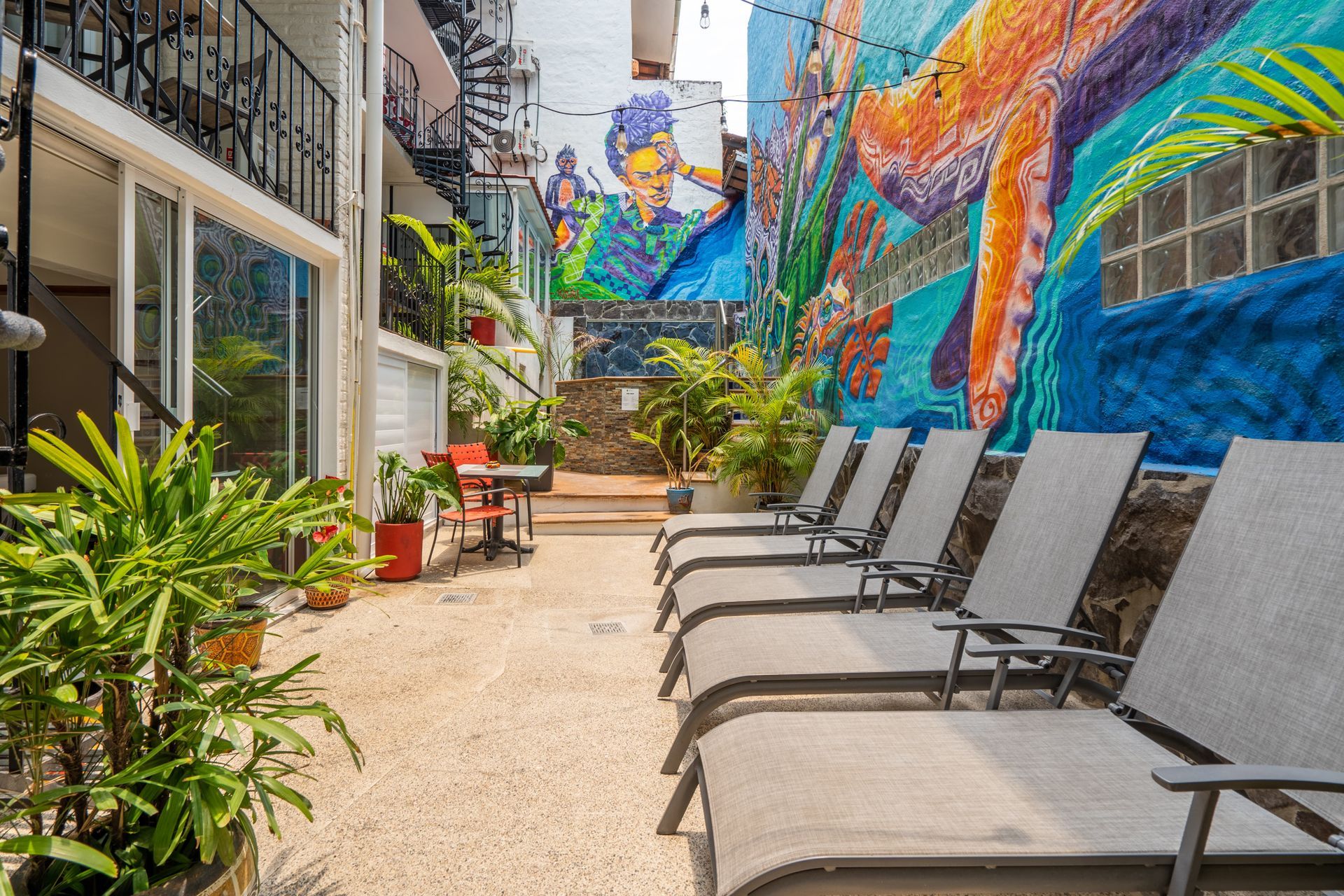 Lounge chairs line a colorful alley with a mural, potted plants, and a balcony in Puerto Vallarta.