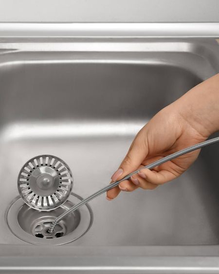Hand using a drain snake to unclog a stainless steel kitchen sink drain