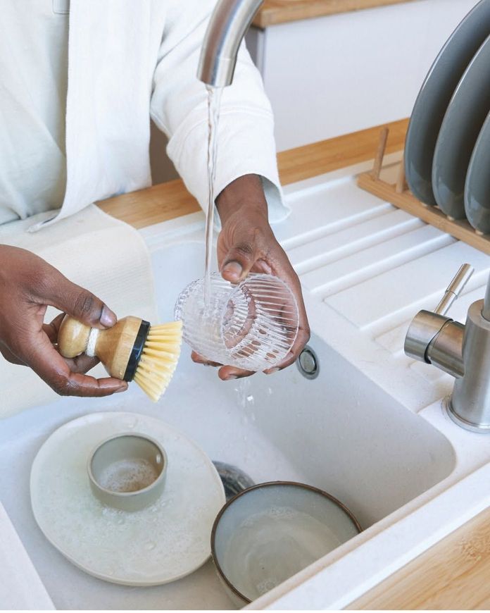 Hands washing dishes in a white kitchen sink with a brush and glass bowl