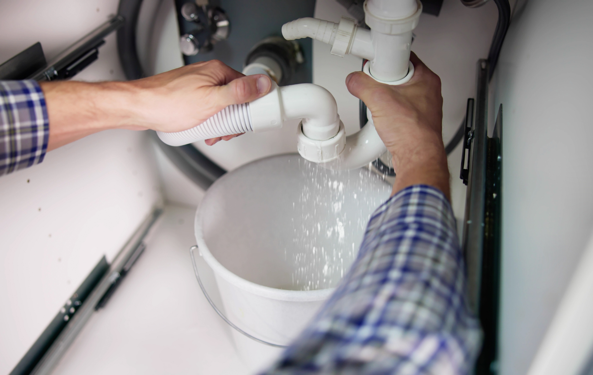 Hands working under a sink, adjusting white plumbing pipes over a bucket as water runs into it