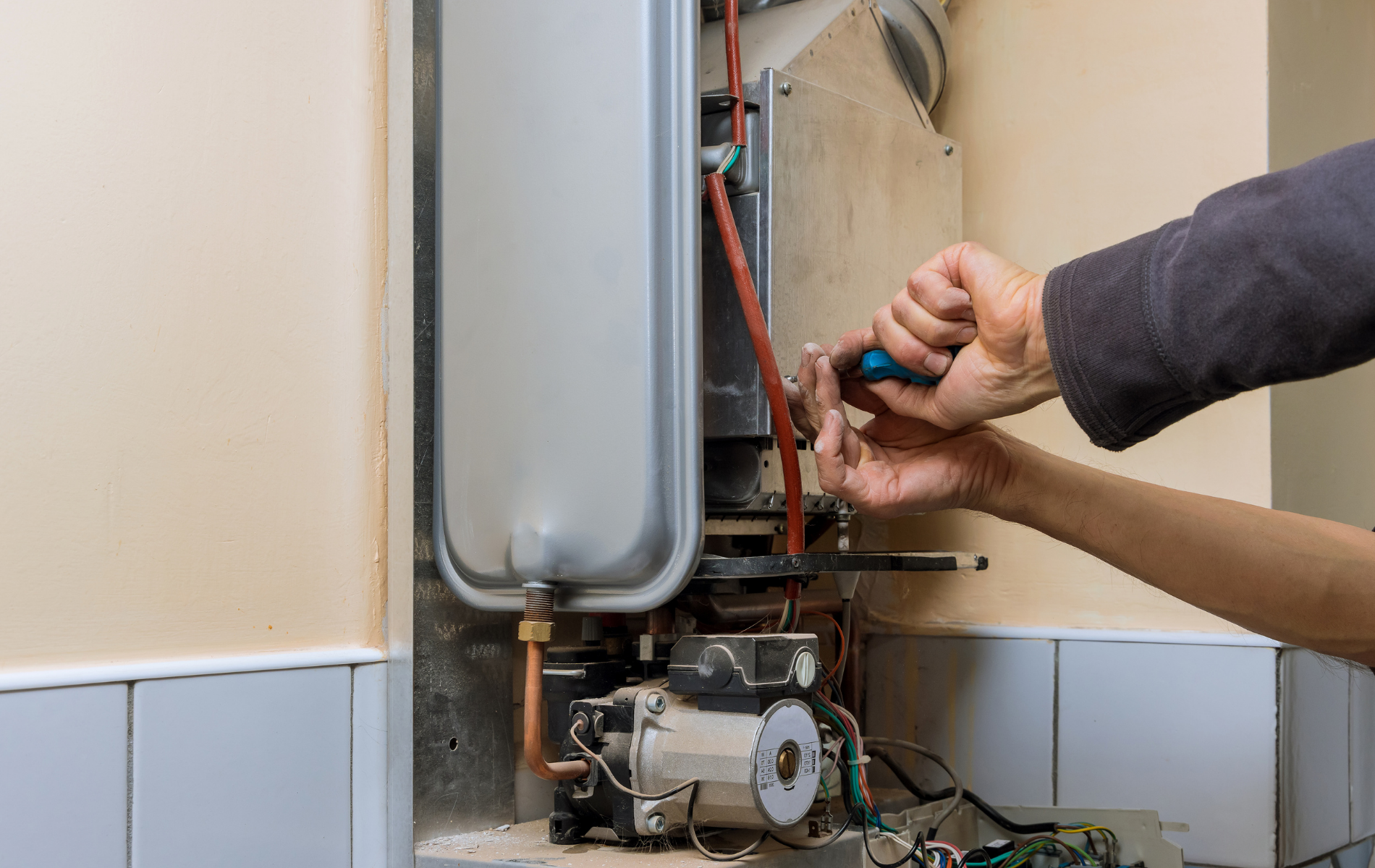 Technician repairing a wall-mounted boiler with a screwdriver, with exposed pipes and wiring.