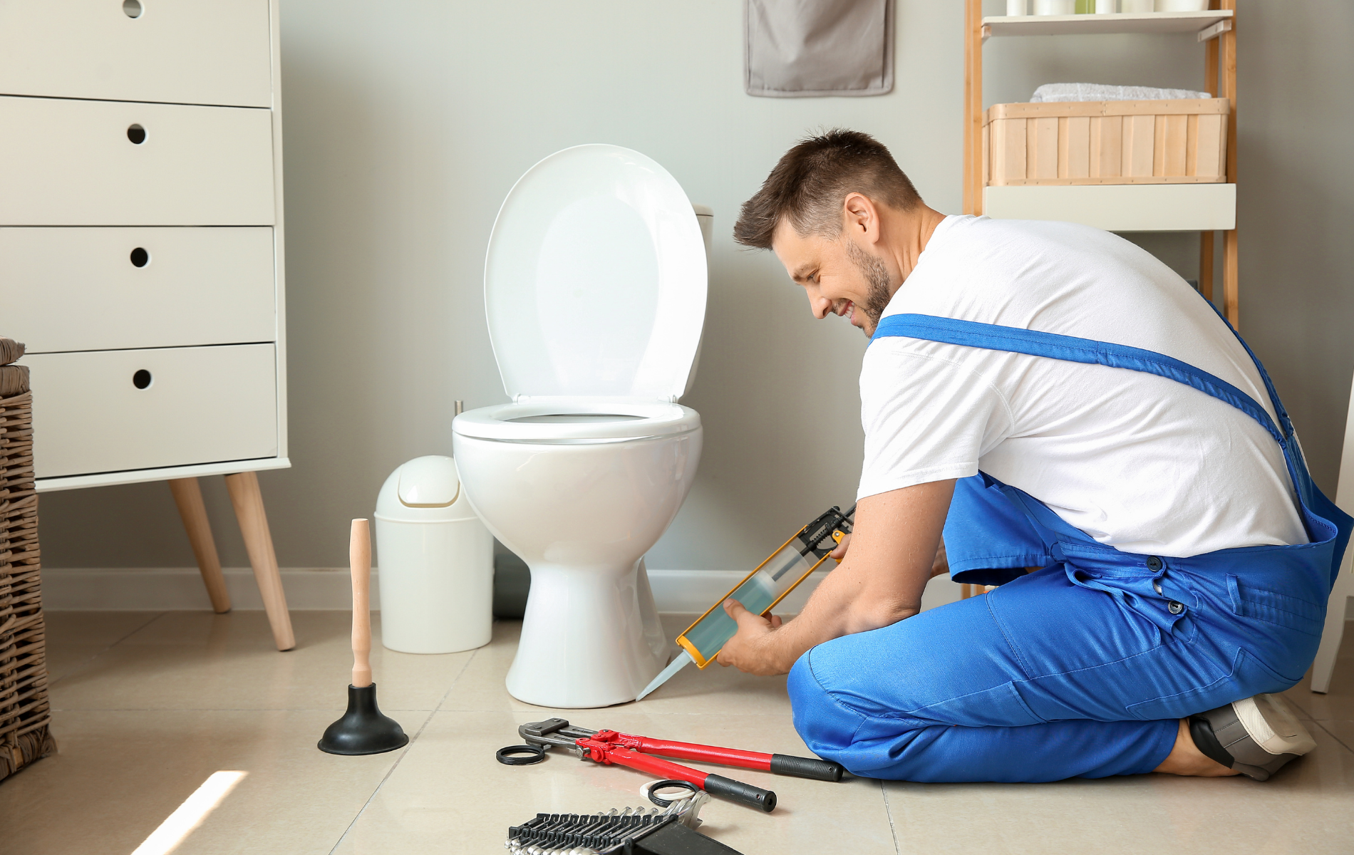 Plumber in blue overalls repairing a toilet in a bathroom, with tools spread on the floor.