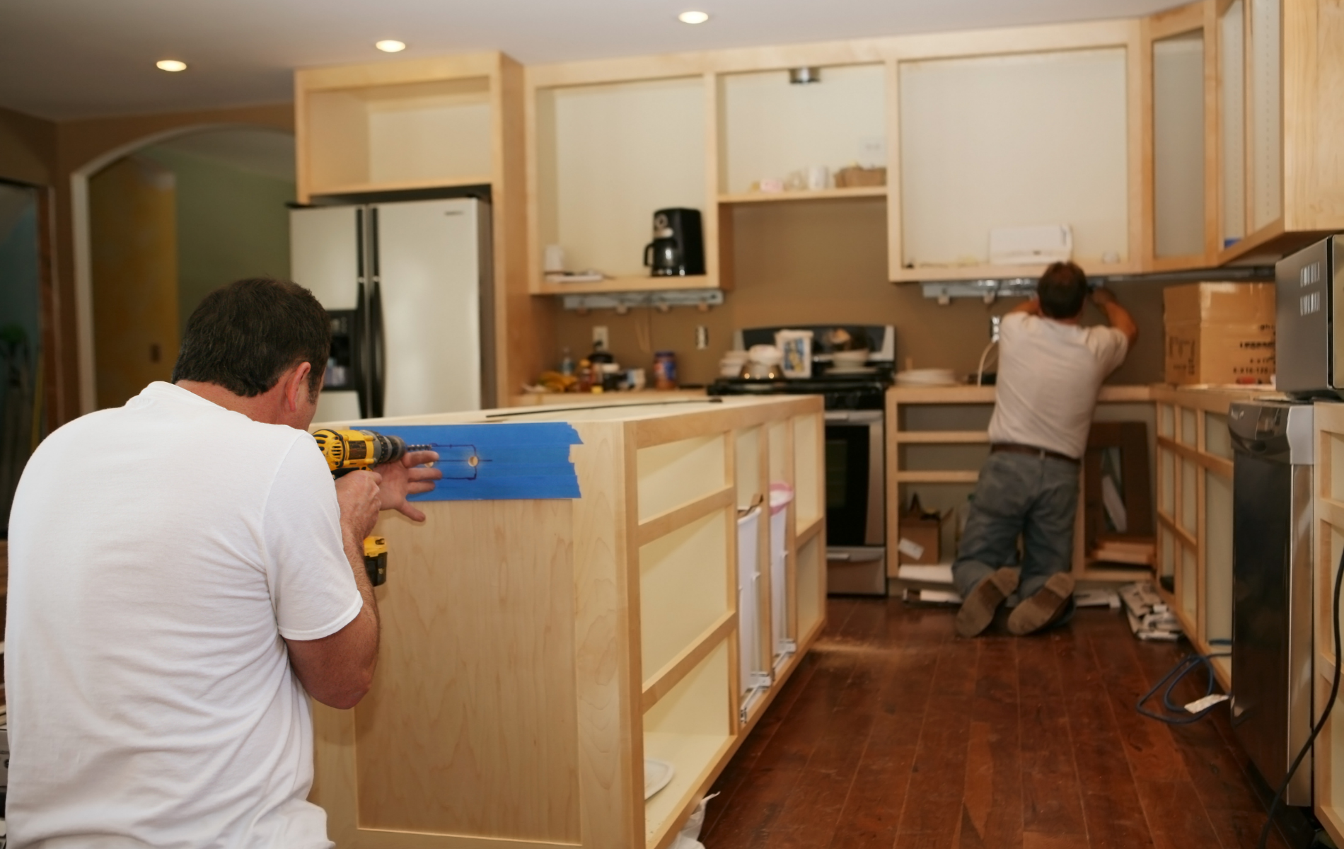Two workers installing kitchen cabinets in a home renovation, with wood flooring and tools visible.