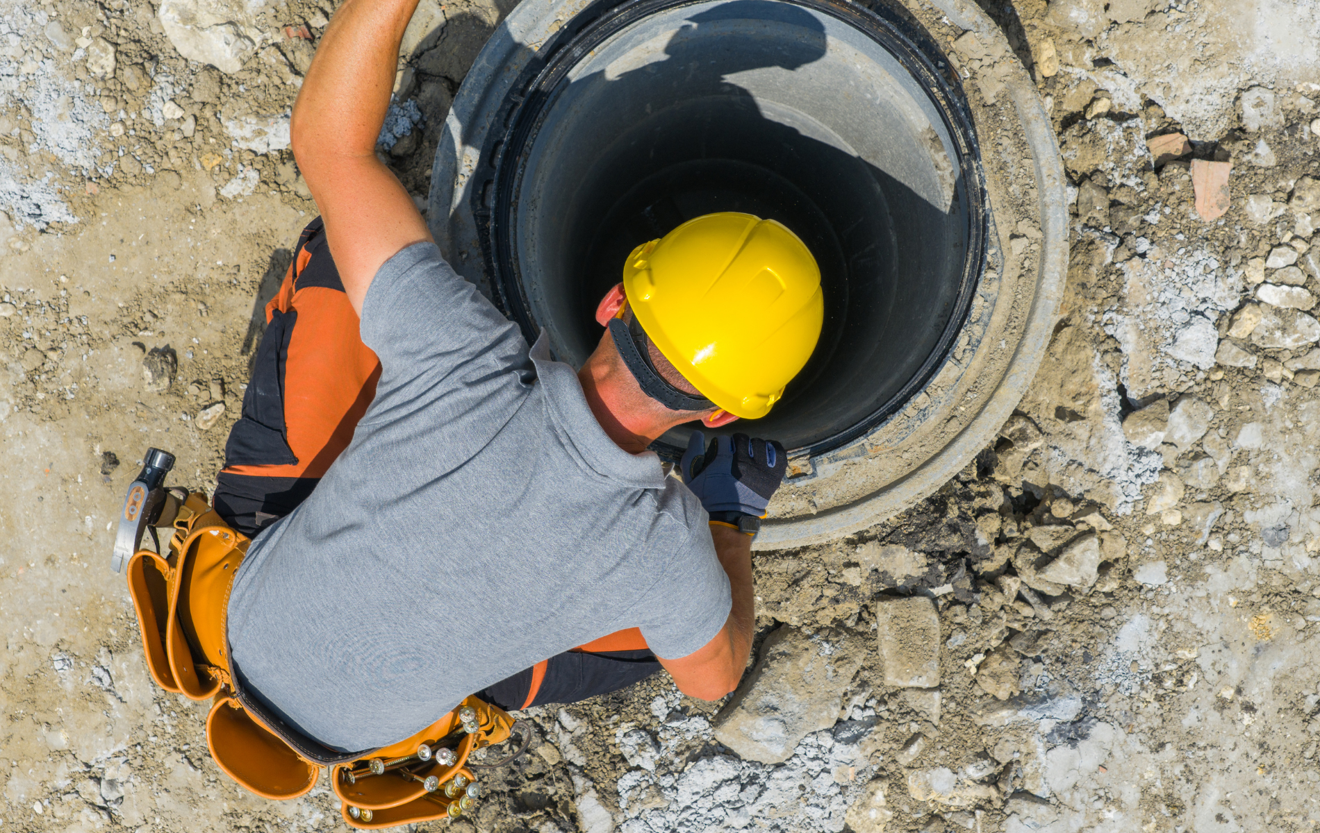 Worker in yellow hard hat descending a dark manhole on a construction site.