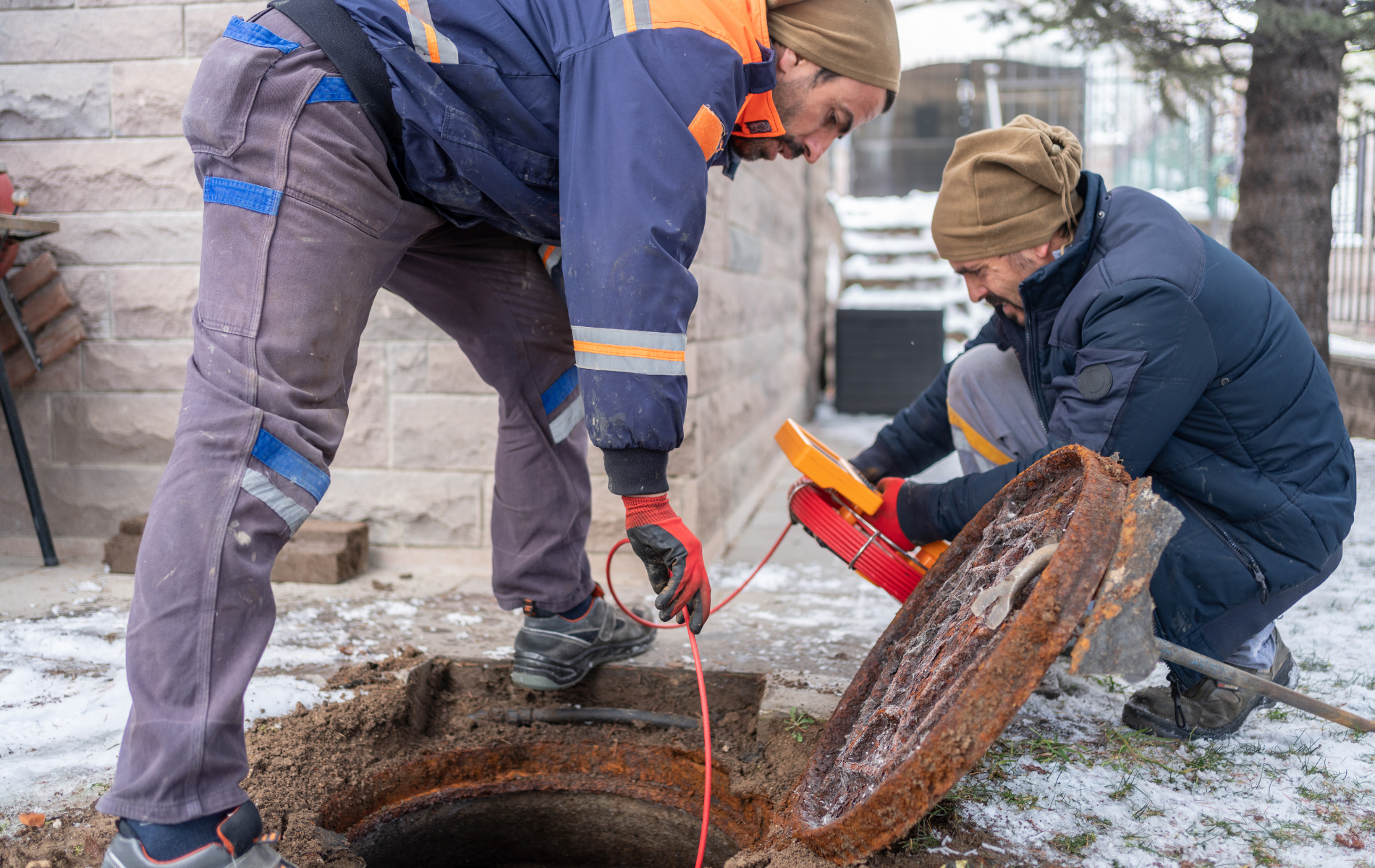 Two workers in winter gear inspect a broken pipe opening outdoors with tools and cables.