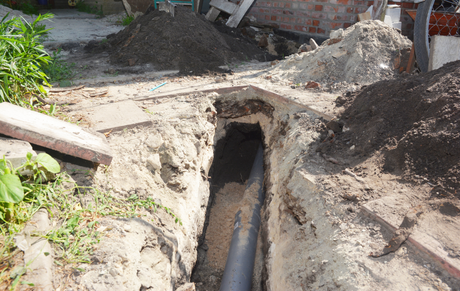 Open trench with exposed pipe beneath a dirt roadway, surrounded by piled soil.