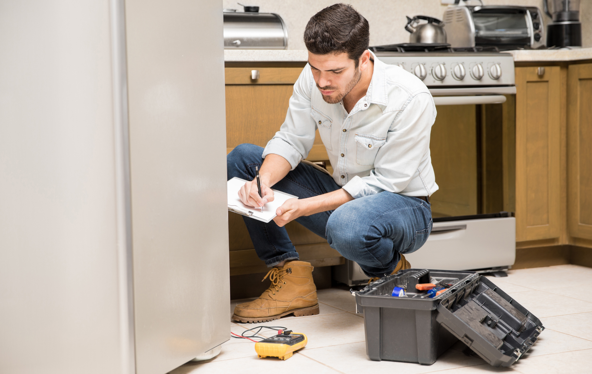 Technician kneeling by a refrigerator in a kitchen, examining tools and open repair equipment on the floor.