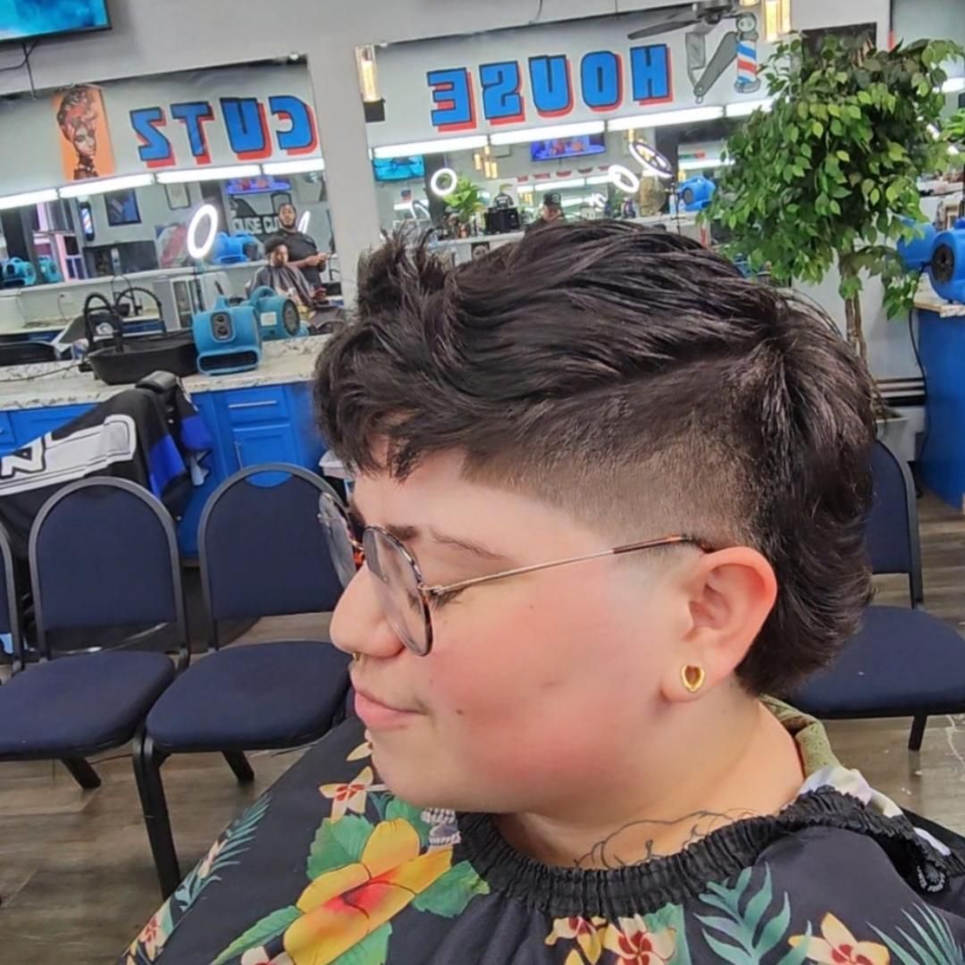 Person with short dark hair and glasses in a barber shop, shown in profile with blue chairs and mirror signage behind.