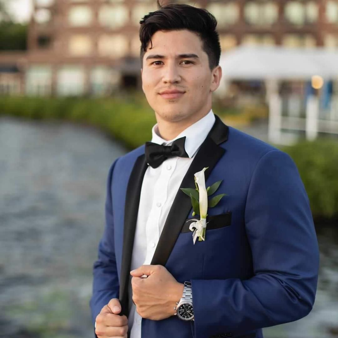 Man in a blue suit and black bow tie standing outdoors with a white boutonniere, blurred building behind him