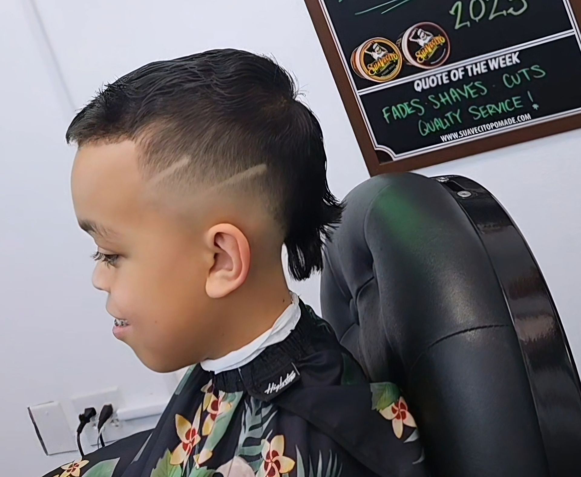 Child with a slicked-back haircut sitting in a barber chair, side profile, looking forward.